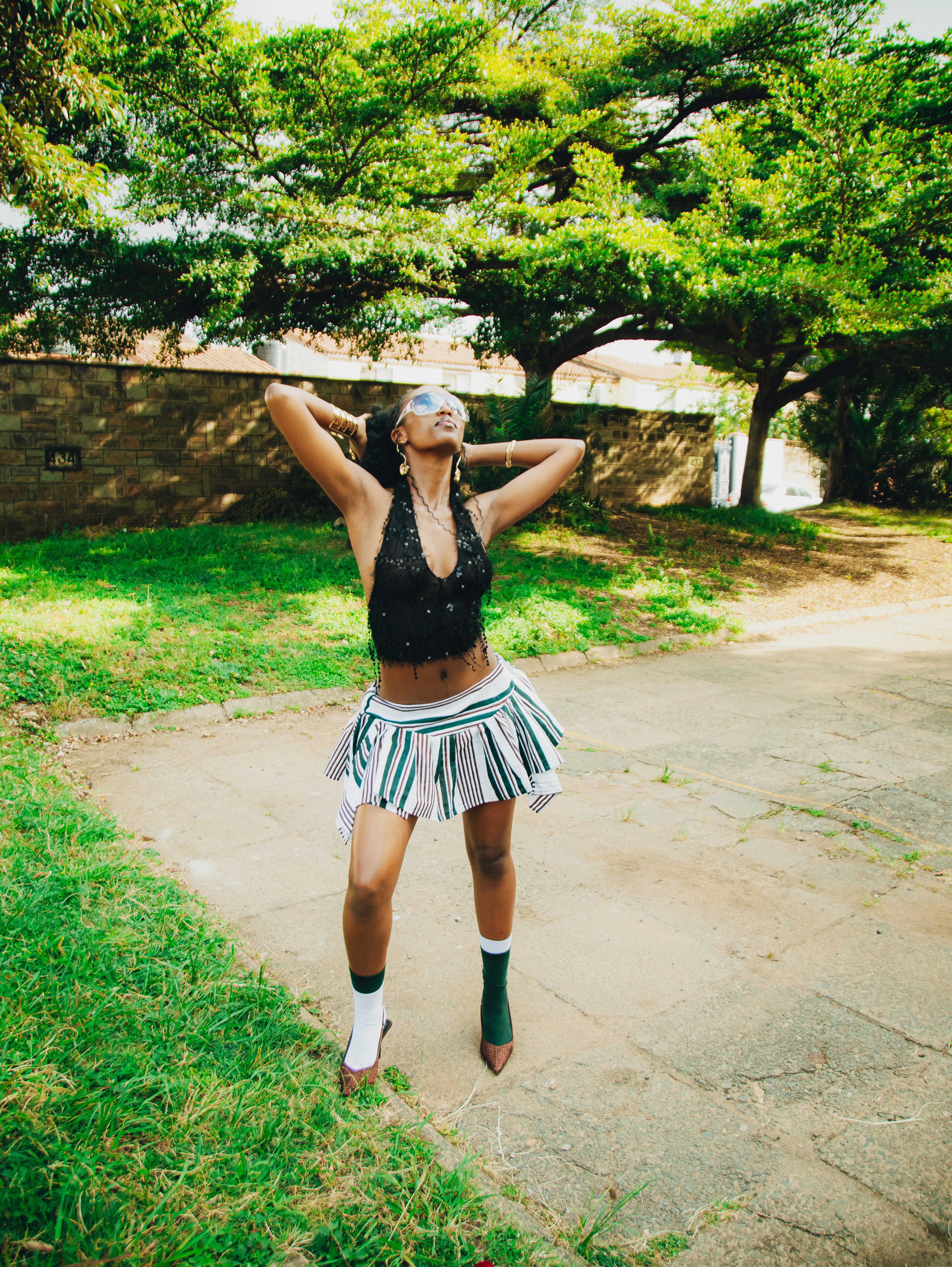 Stylish woman in a striped skirt posing under green trees.