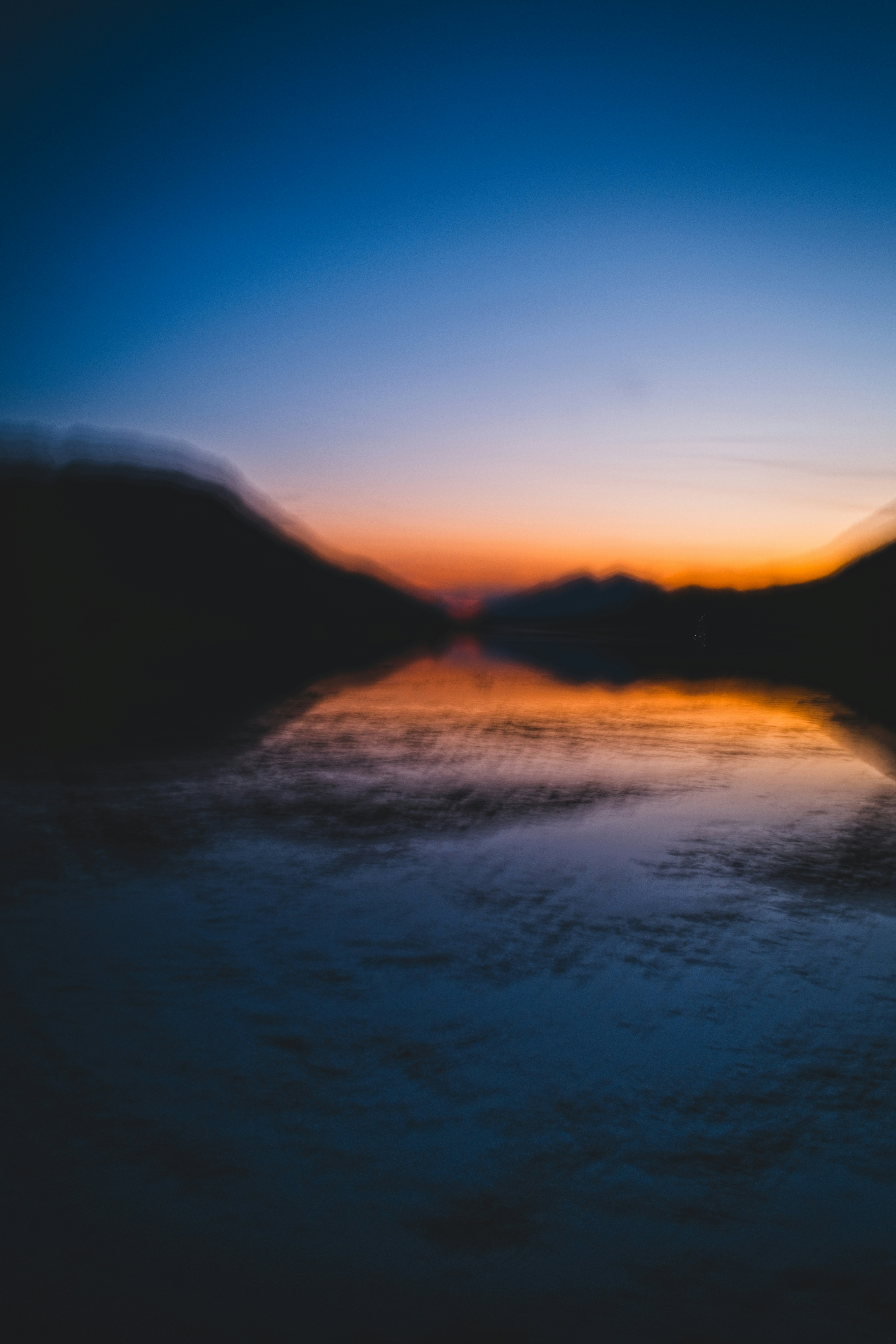 Pôr do sol laranja e azul refletido na água com montanhas escuras.