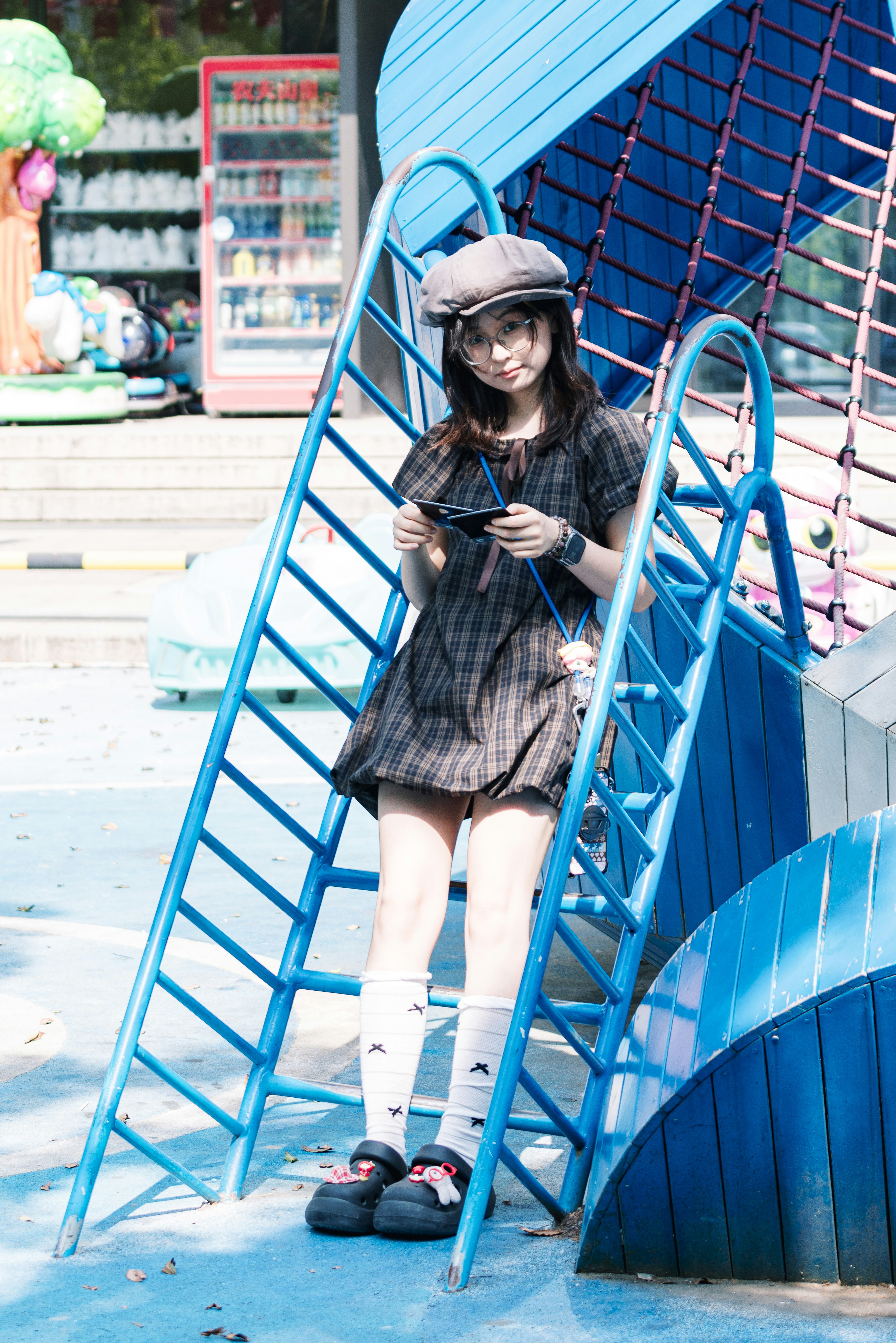 Stylish young woman on a blue playground ladder holding sunglasses.