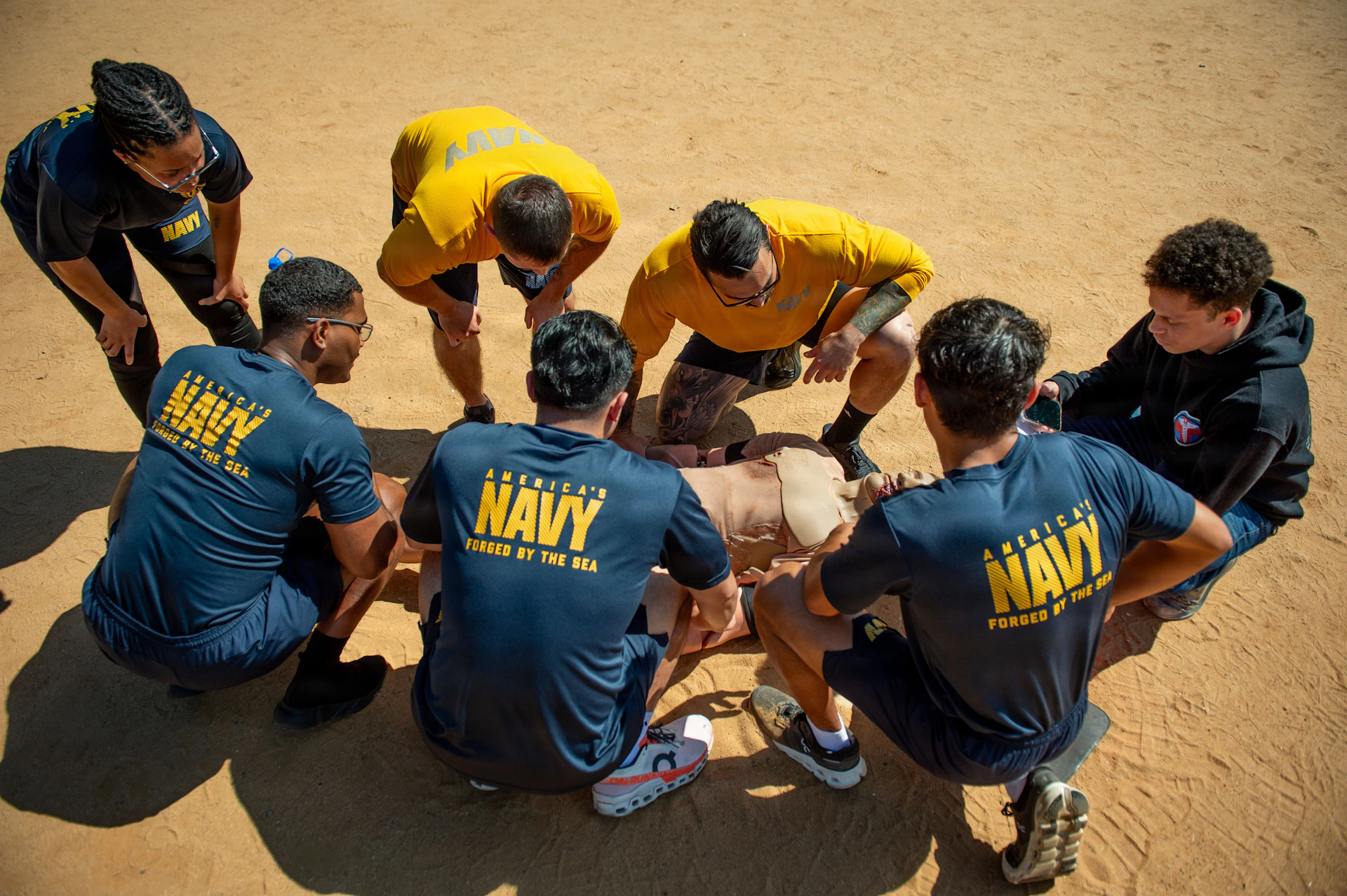 Navy personnel train around a dummy on sandy ground.