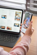 Hand holding smartphone displaying content, with tablet and keyboard.