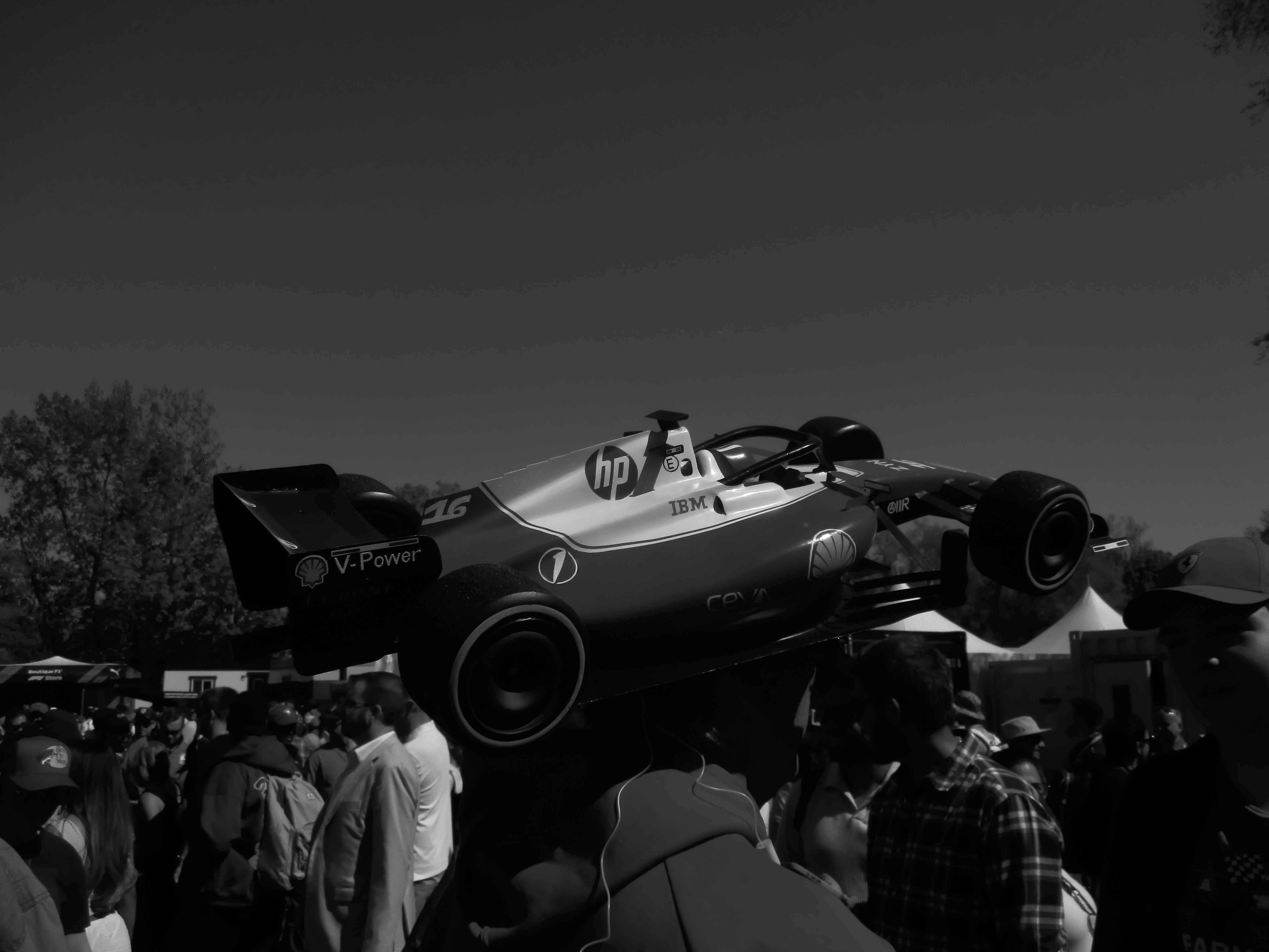 Low-angle shot of a Formula-style race car lifted above a dense crowd at an outdoor event; the scene emphasizes the engineering display and spectator interest.