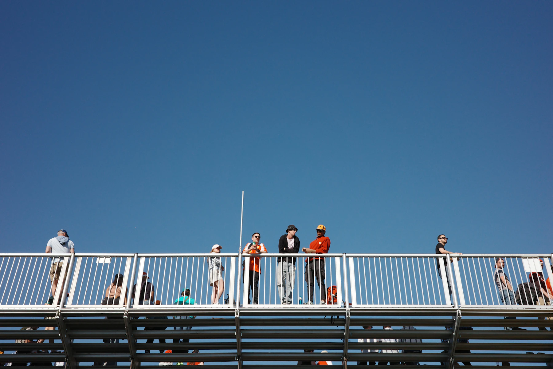Spectators standing along the top rail of a set of metal bleachers, seen from below against a deep blue sky
