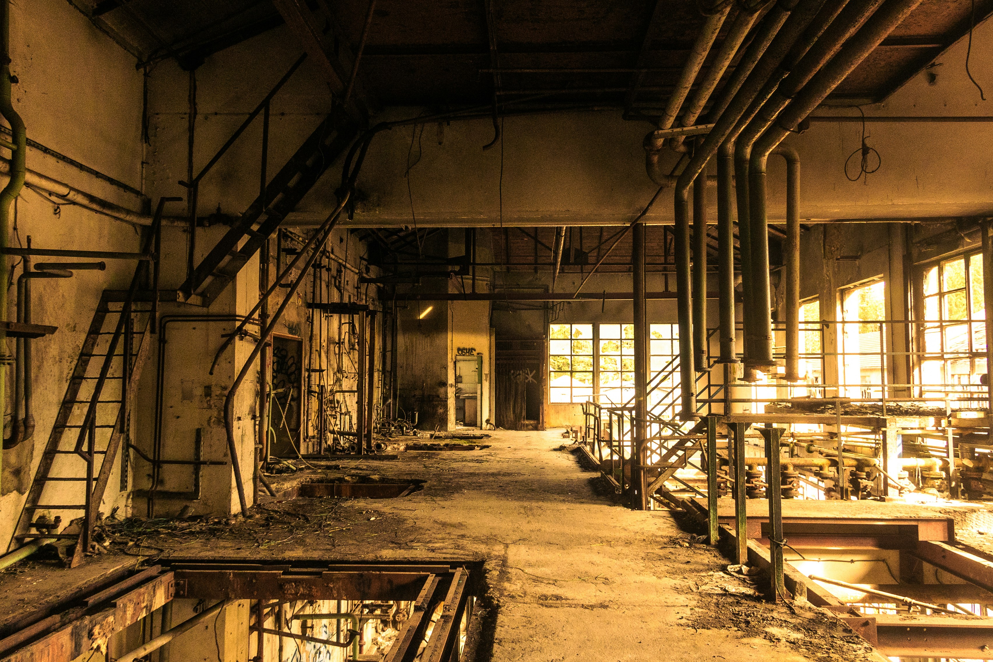 An abandoned, decaying industrial factory interior with pipes and windows.