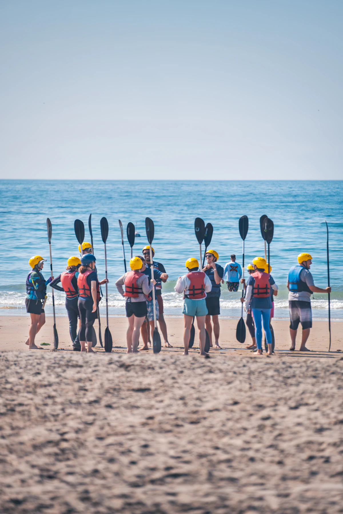 A team in life vests and helmets holding kayak paddles on a beach before a paddling outing