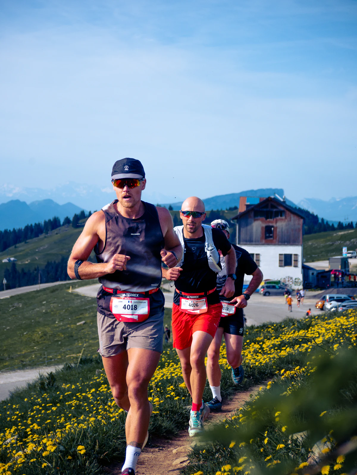 Dos corredores de trail suben un sendero de montaña entre flores silvestres amarillas, con picos alpinos al fondo