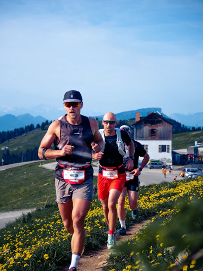 Dos corredores de trail suben un sendero de montaña entre flores silvestres amarillas, con picos alpinos al fondo
