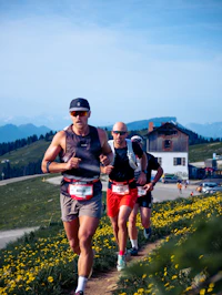 Dos corredores de trail suben un sendero de montaña entre flores silvestres amarillas, con picos alpinos al fondo