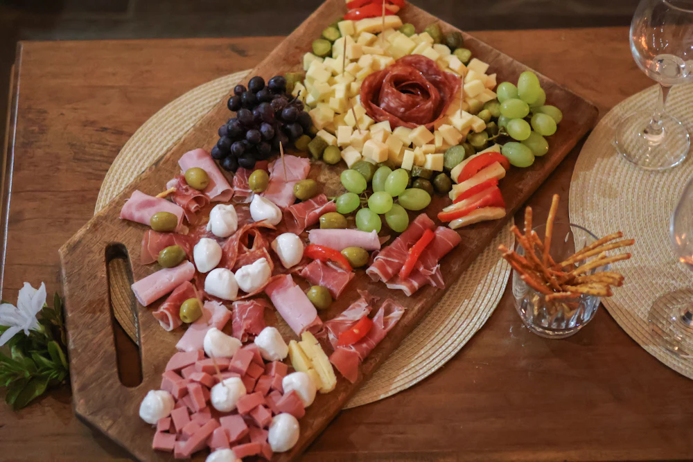 A wooden board of cheese cubes, rolled cured meats, bocconcini, grapes and breadsticks in a glass beside it