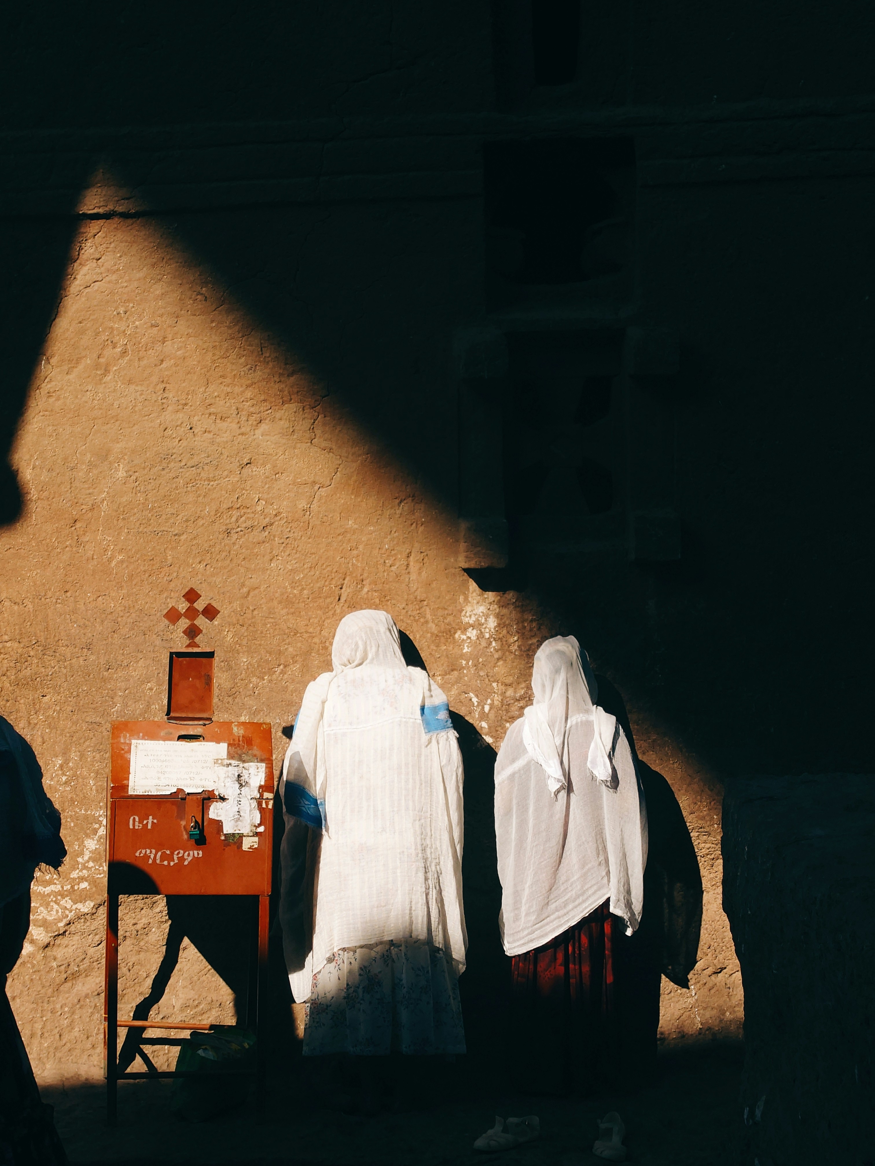 Two robed women stand near a cross in sunlight.