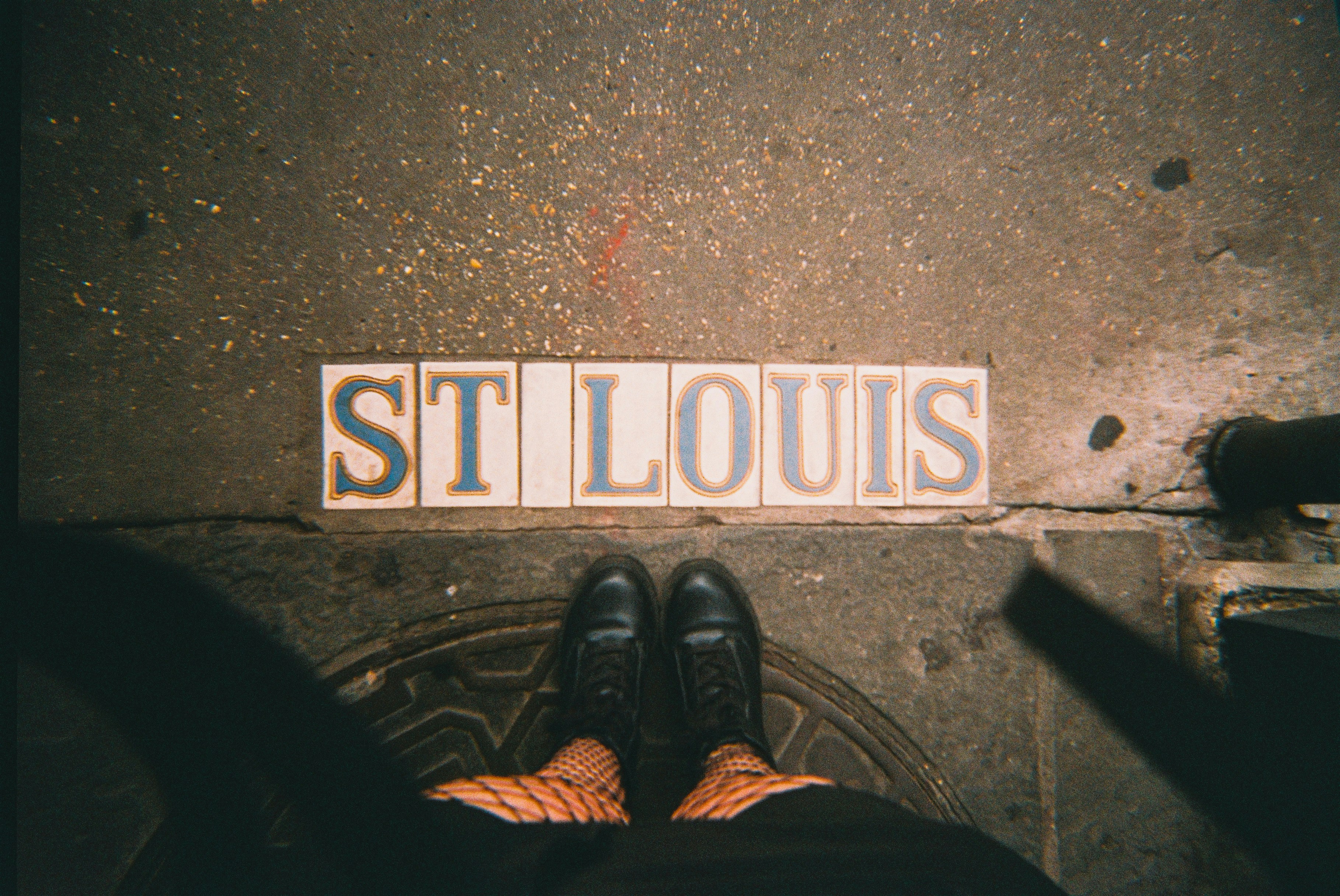 Feet standing on "st louis" tiles on a concrete sidewalk.