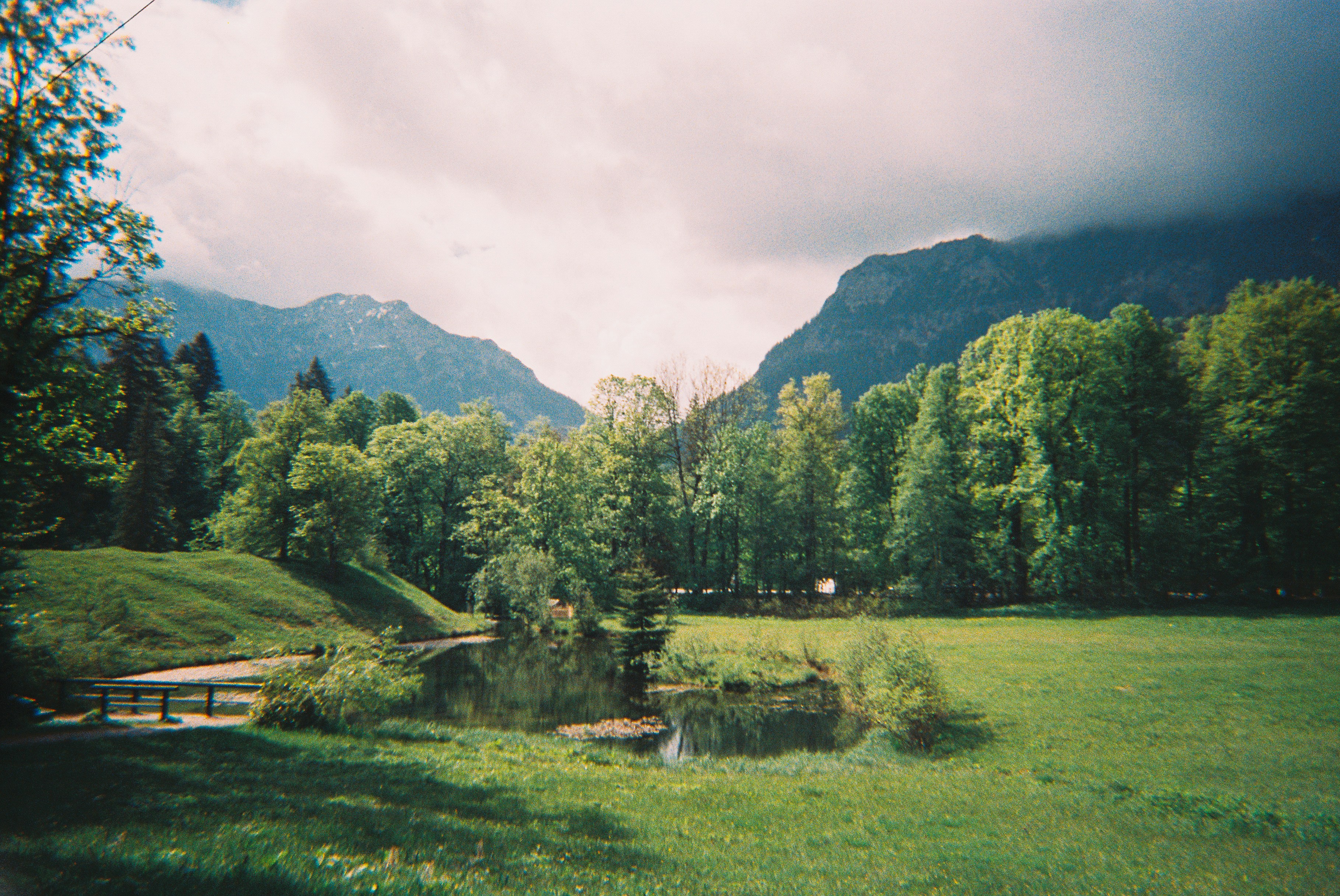 Green field, lush trees, and mountains under a cloudy sky.