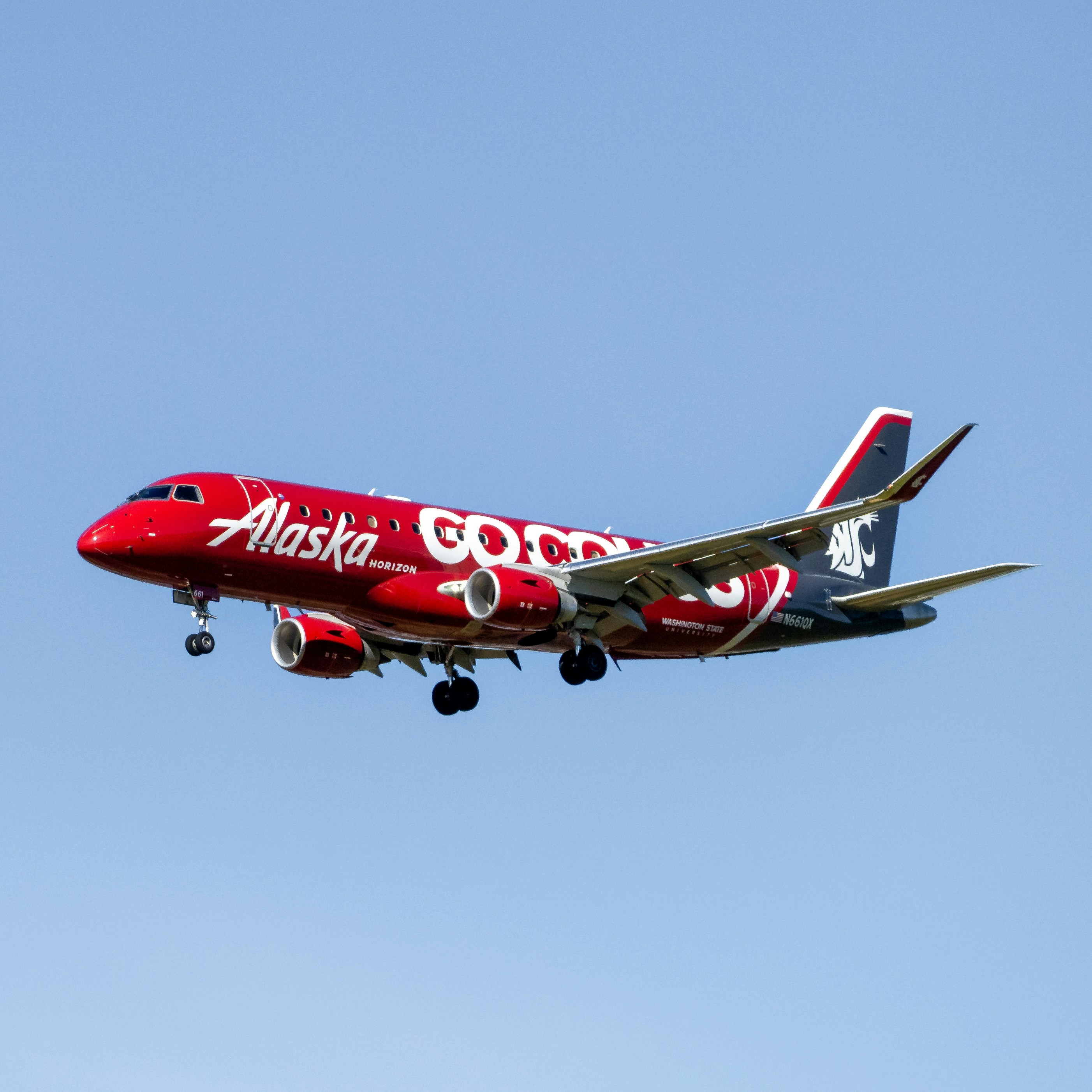 Red Alaska Airlines jet approaches for landing against a pale blue sky, with landing gear extended and engines visible; image clearly captures branding and aircraft details.