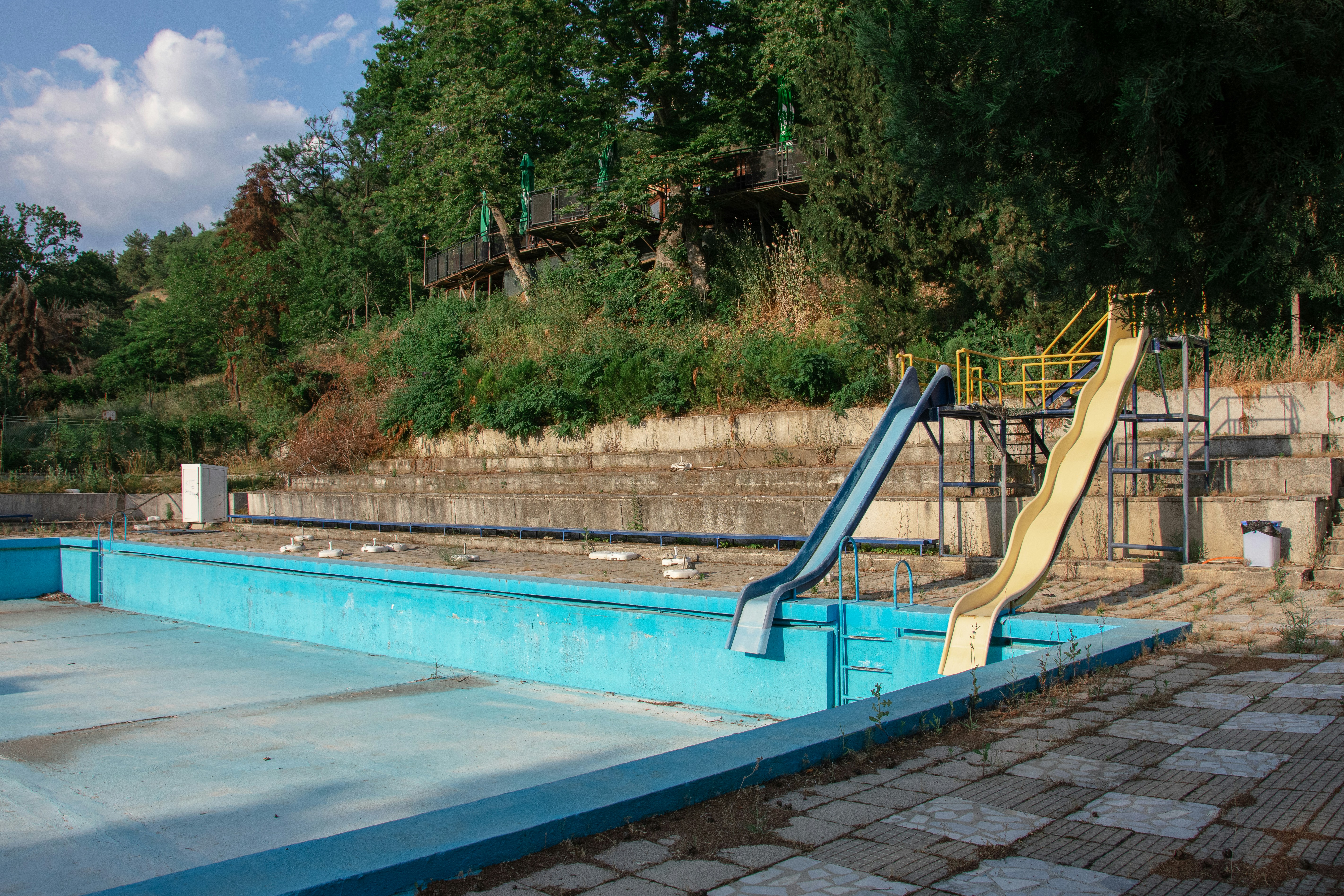 Abandoned swimming pool with two slides and overgrown vegetation