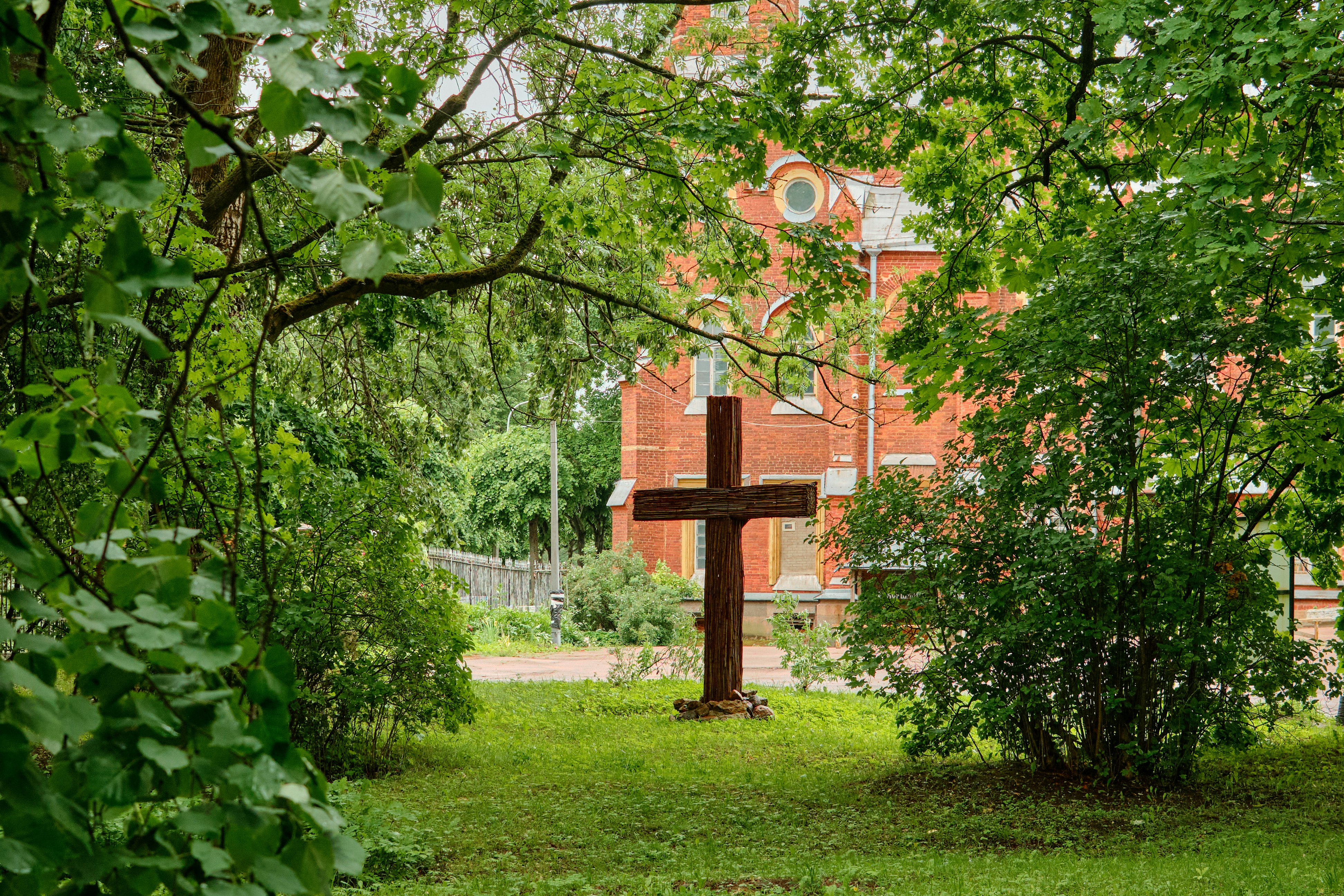 Wooden cross surrounded by green trees with a red building.