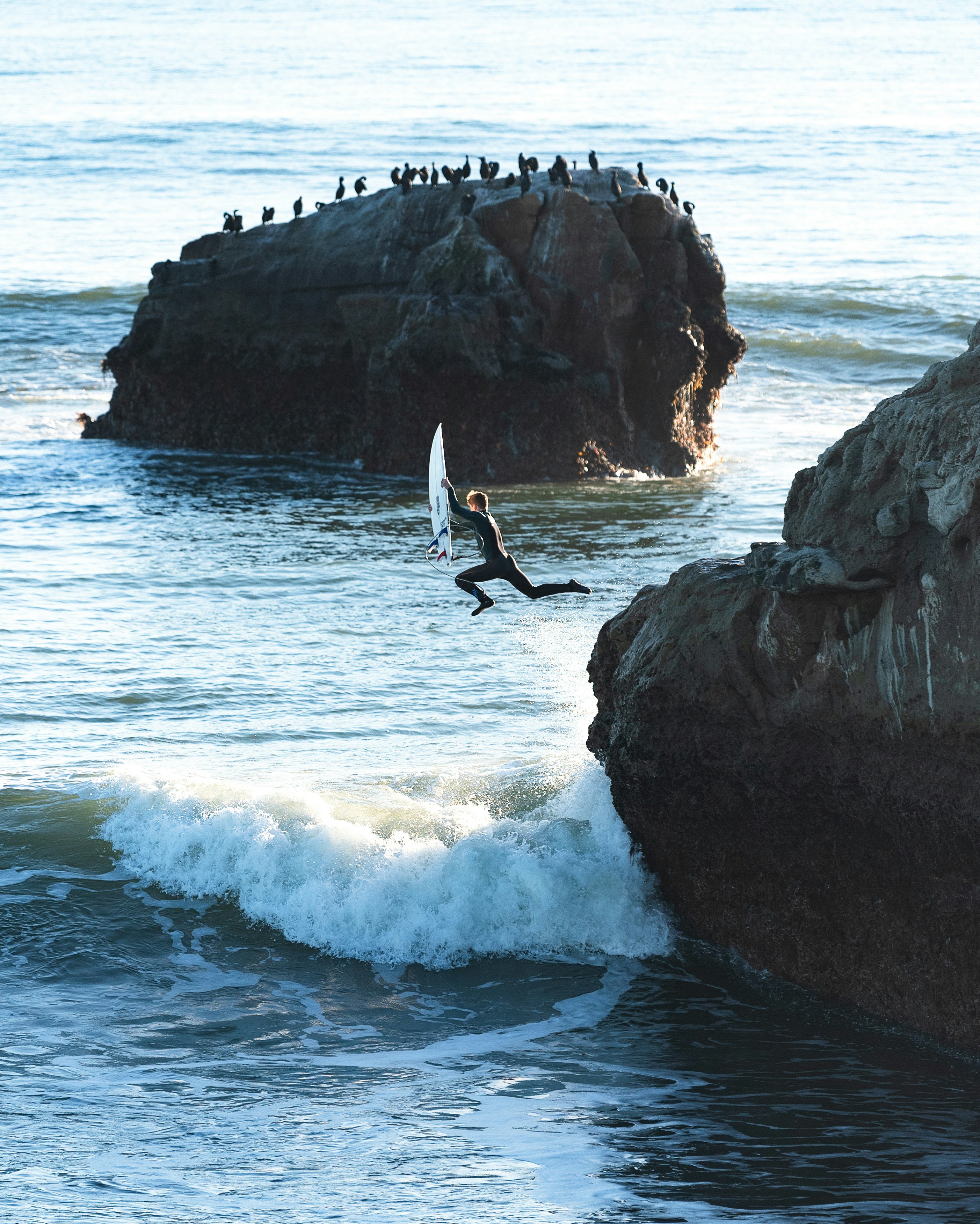 Surfer jumping from a rock into ocean with surfboard.