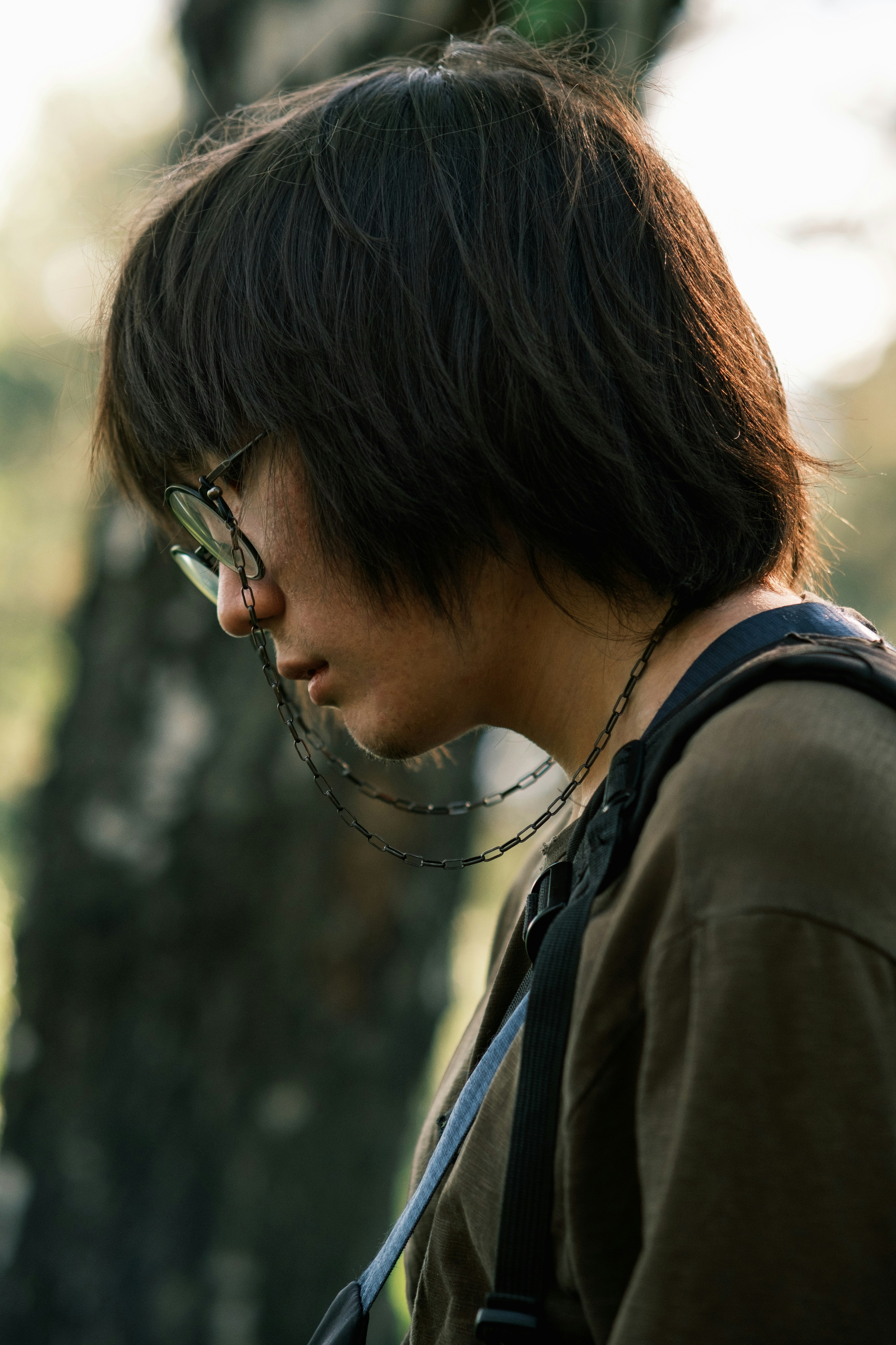A person with dark hair and glasses looks down outdoors.