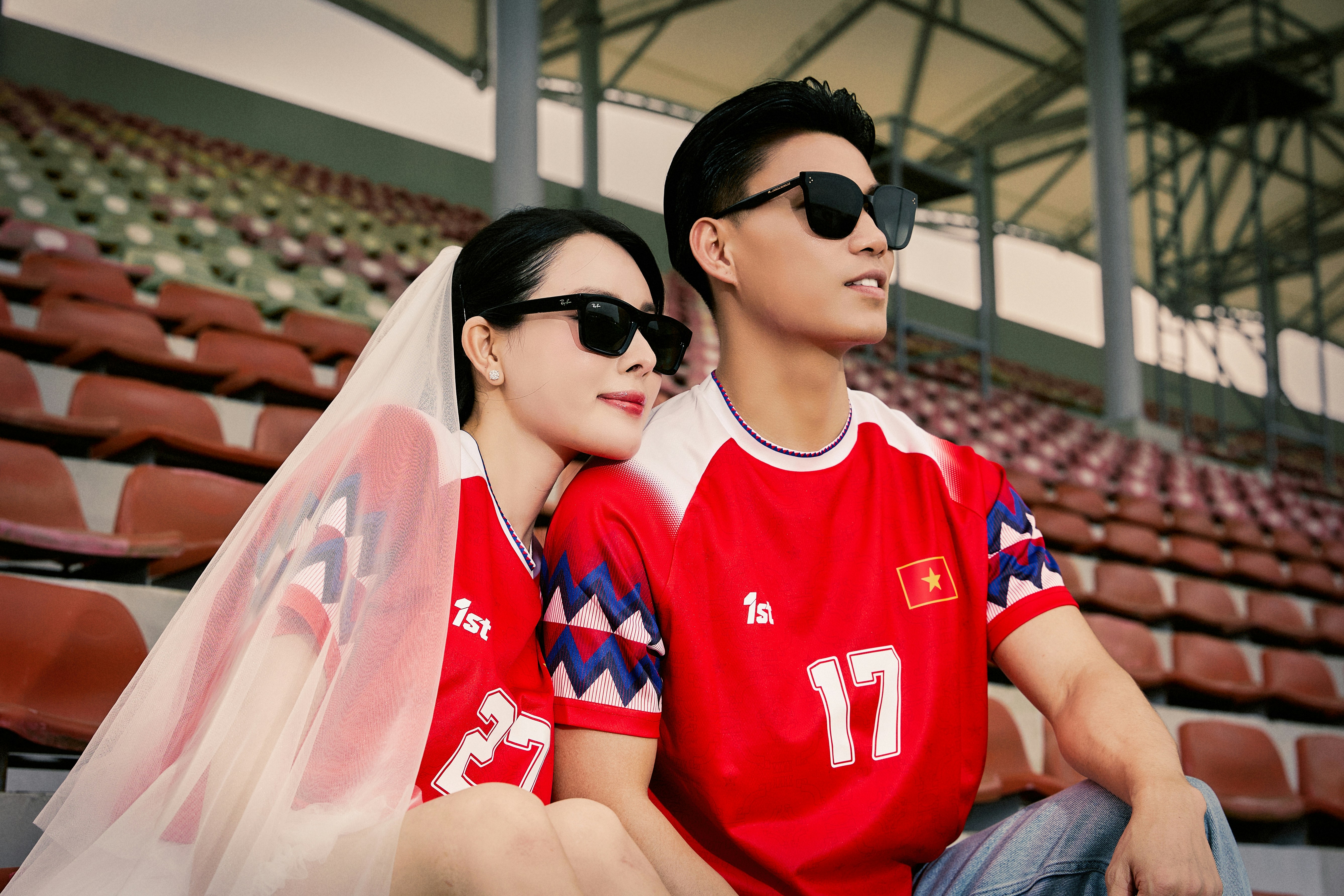 Pasangan mengenakan kaus merah, kacamata hitam, dan kerudung di stadion.