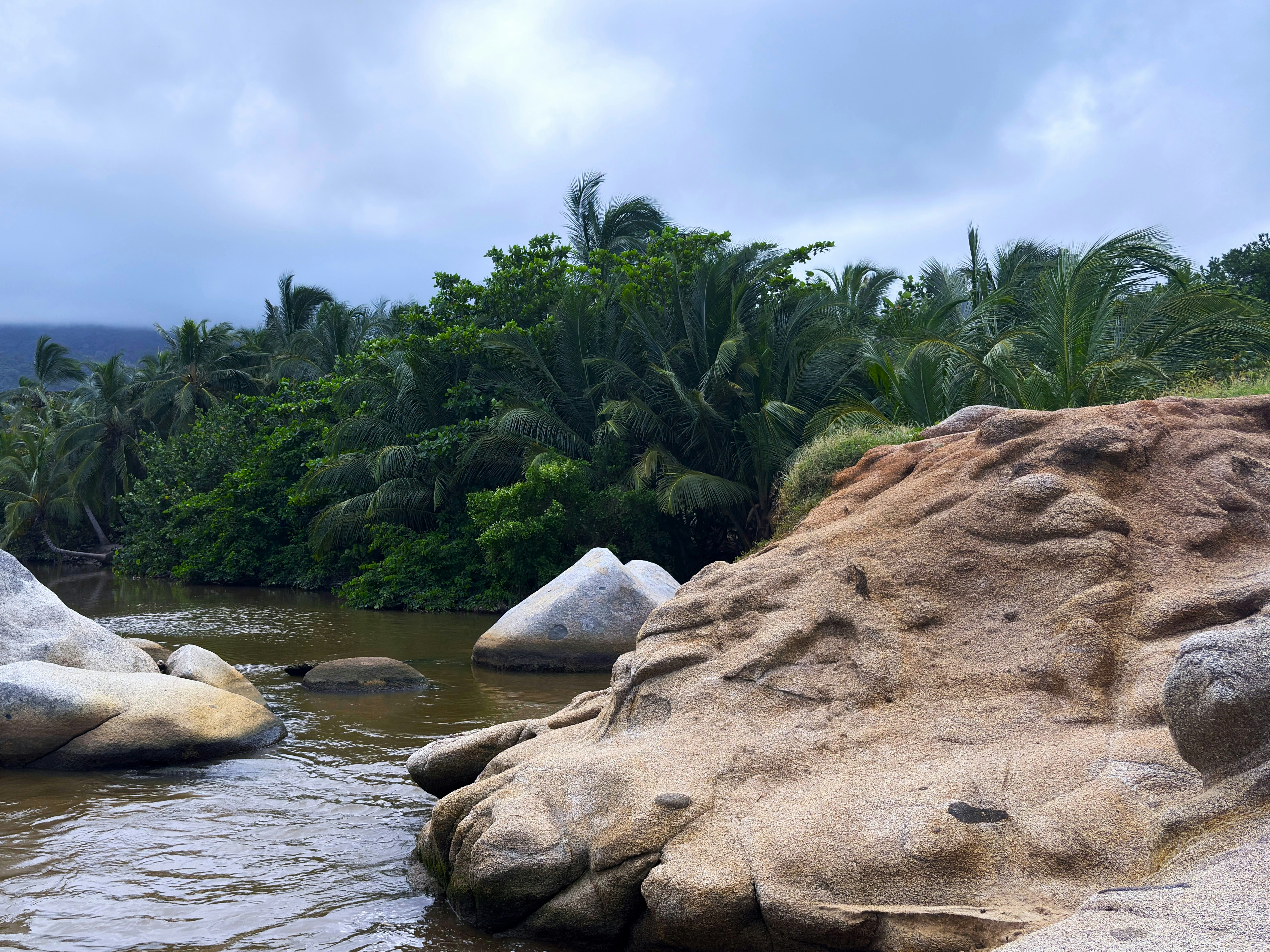 Tropical river flows past large rocks under a cloudy sky.