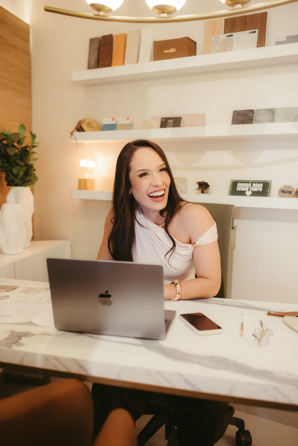 A wellness practitioner laughing at her studio desk with a laptop and phone, product samples on shelves behind her