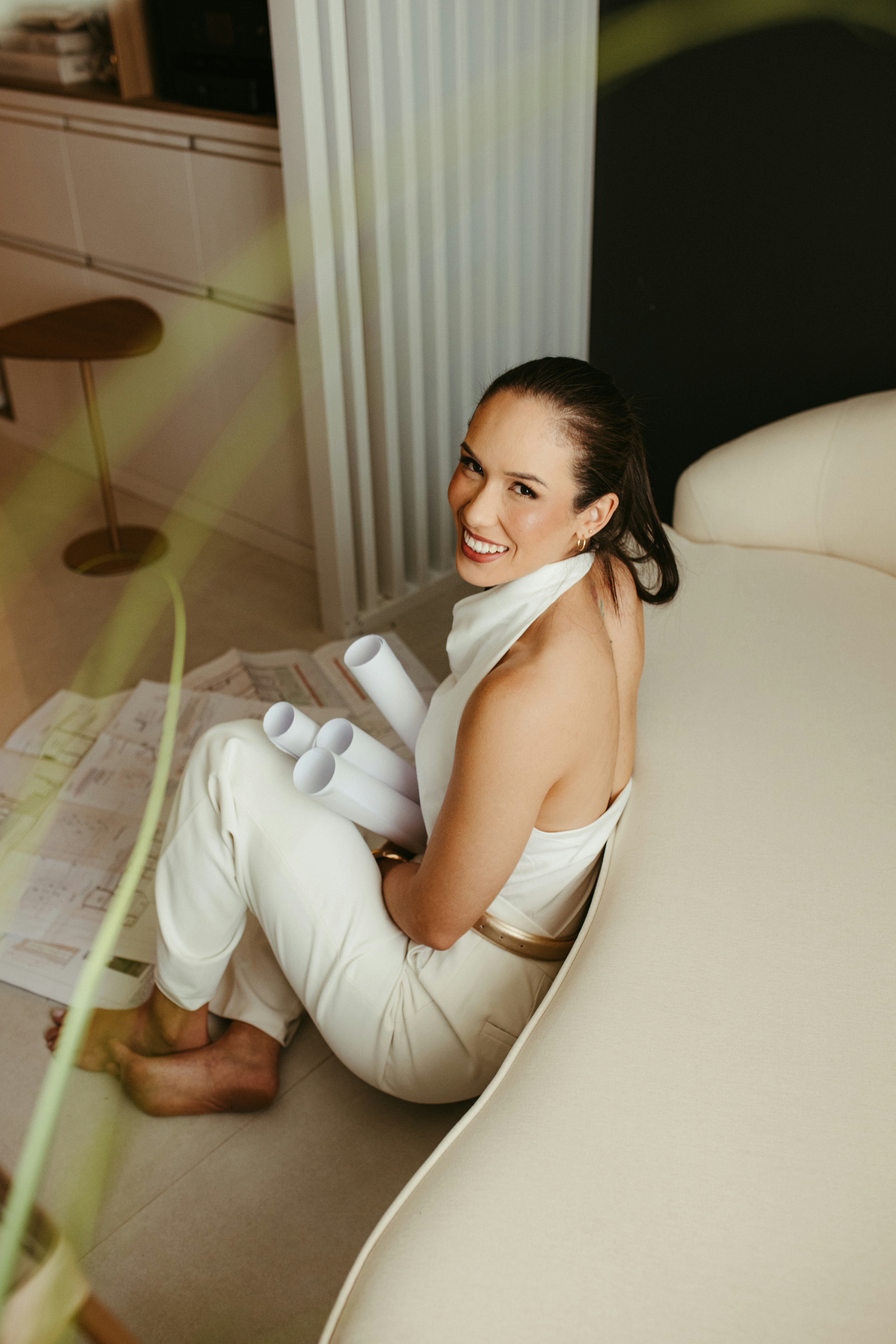 A smiling woman holding blueprints sits on the floor.