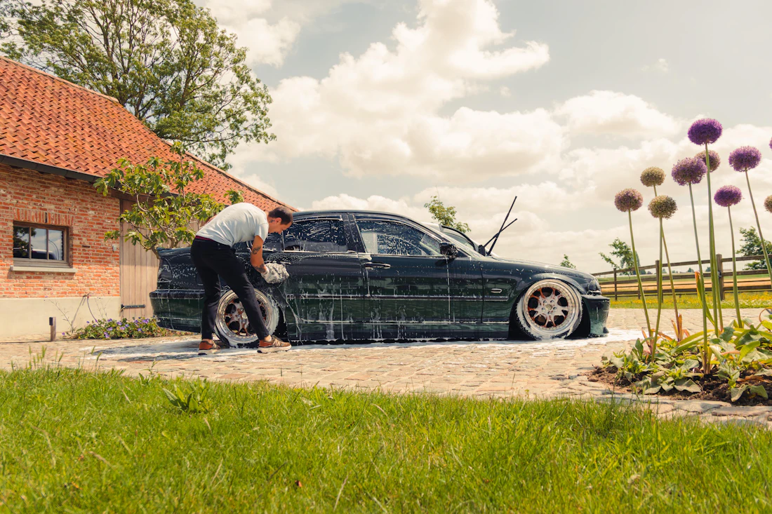 A man washing a dark green car in a sunny driveway