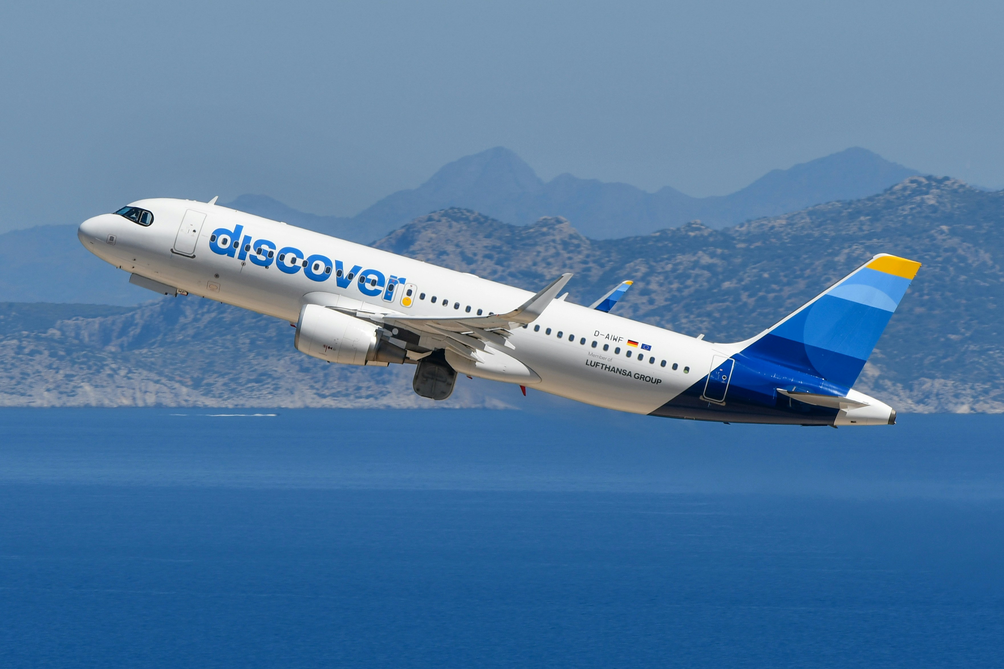Discover airplane ascending over deep blue sea and distant mountains.