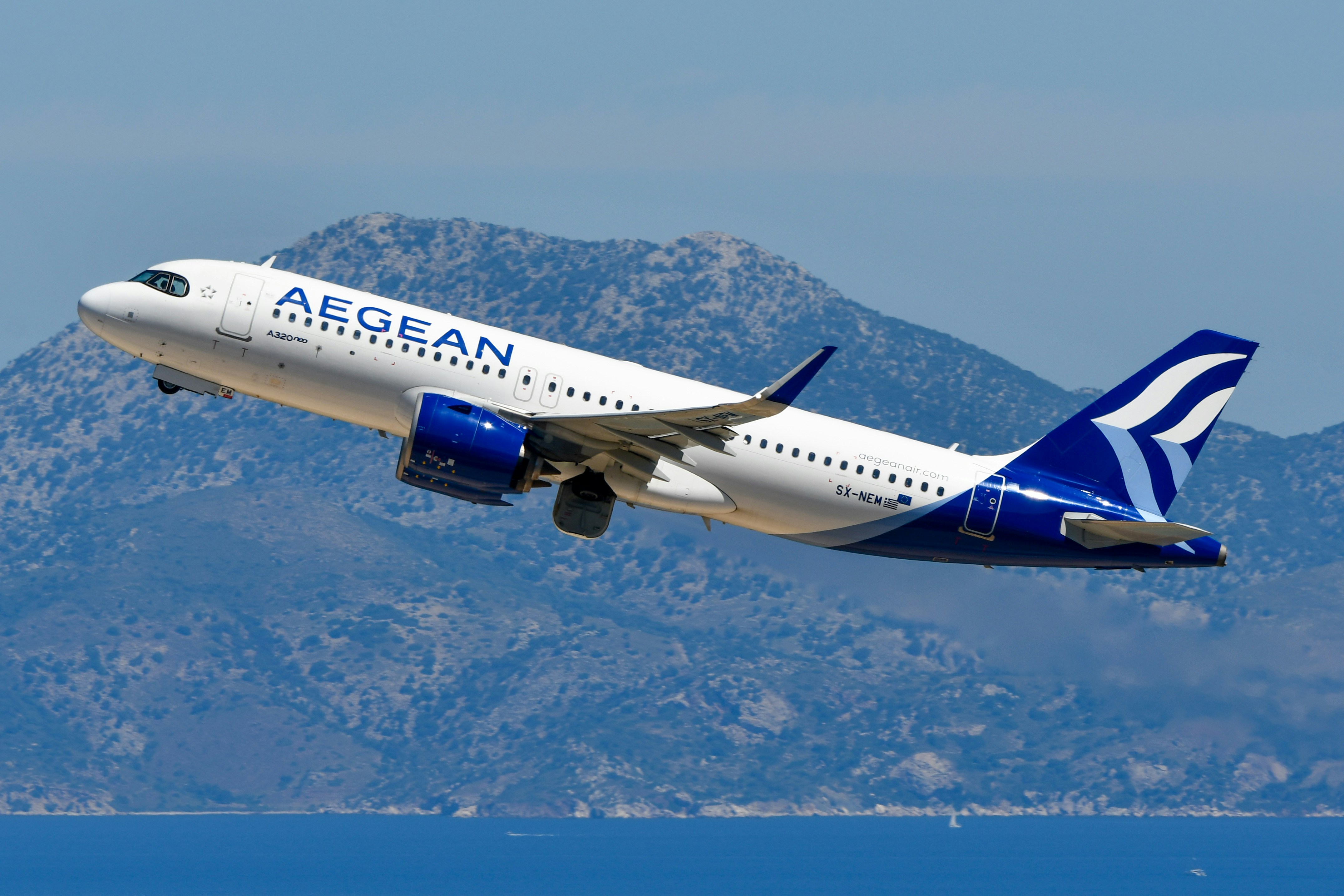 An aegean airlines plane takes off over water with mountains.