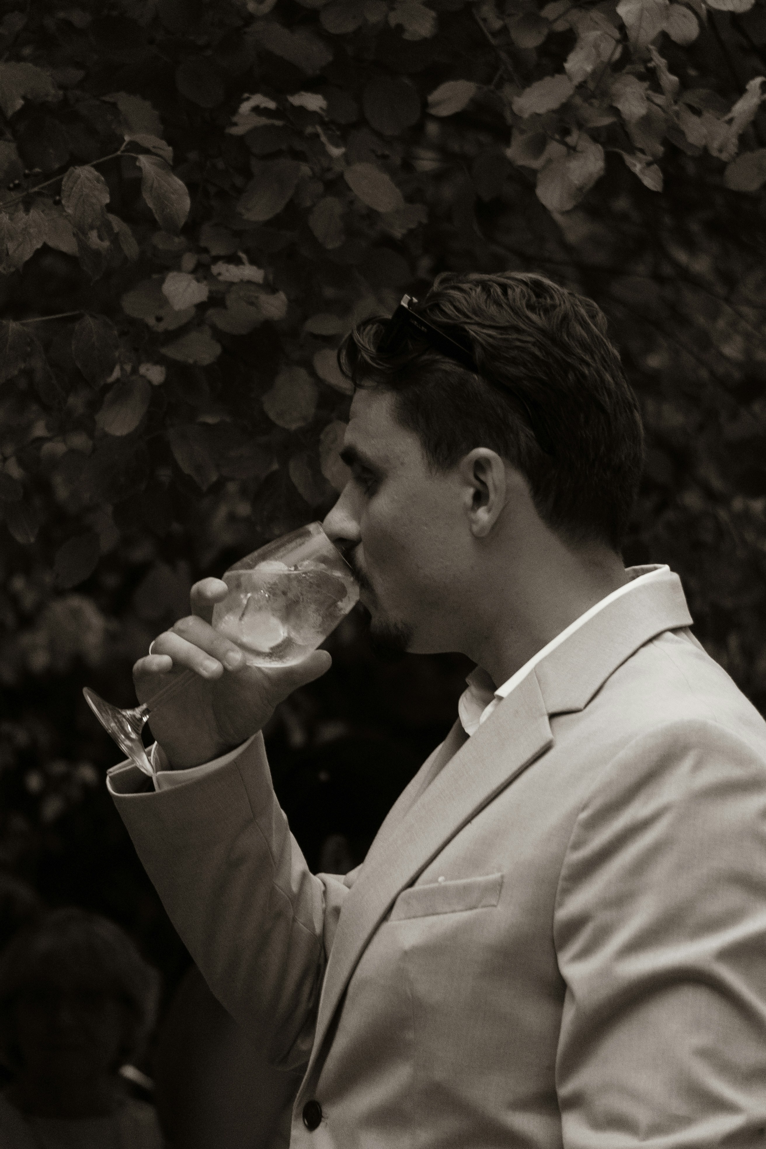 Man in a suit drinking from a glass against foliage