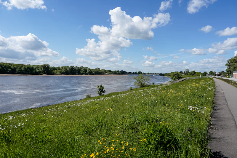 River with a green bank and a paved path under a cloudy blue sky