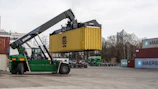 A green reach stacker lifting a yellow shipping container.