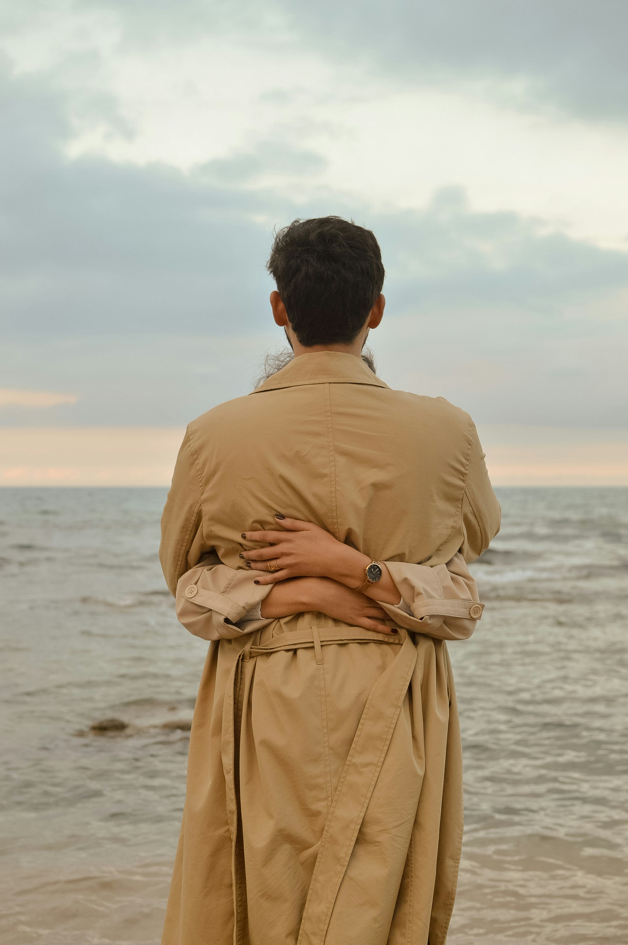 Person in trench coat embraced by another by the ocean.