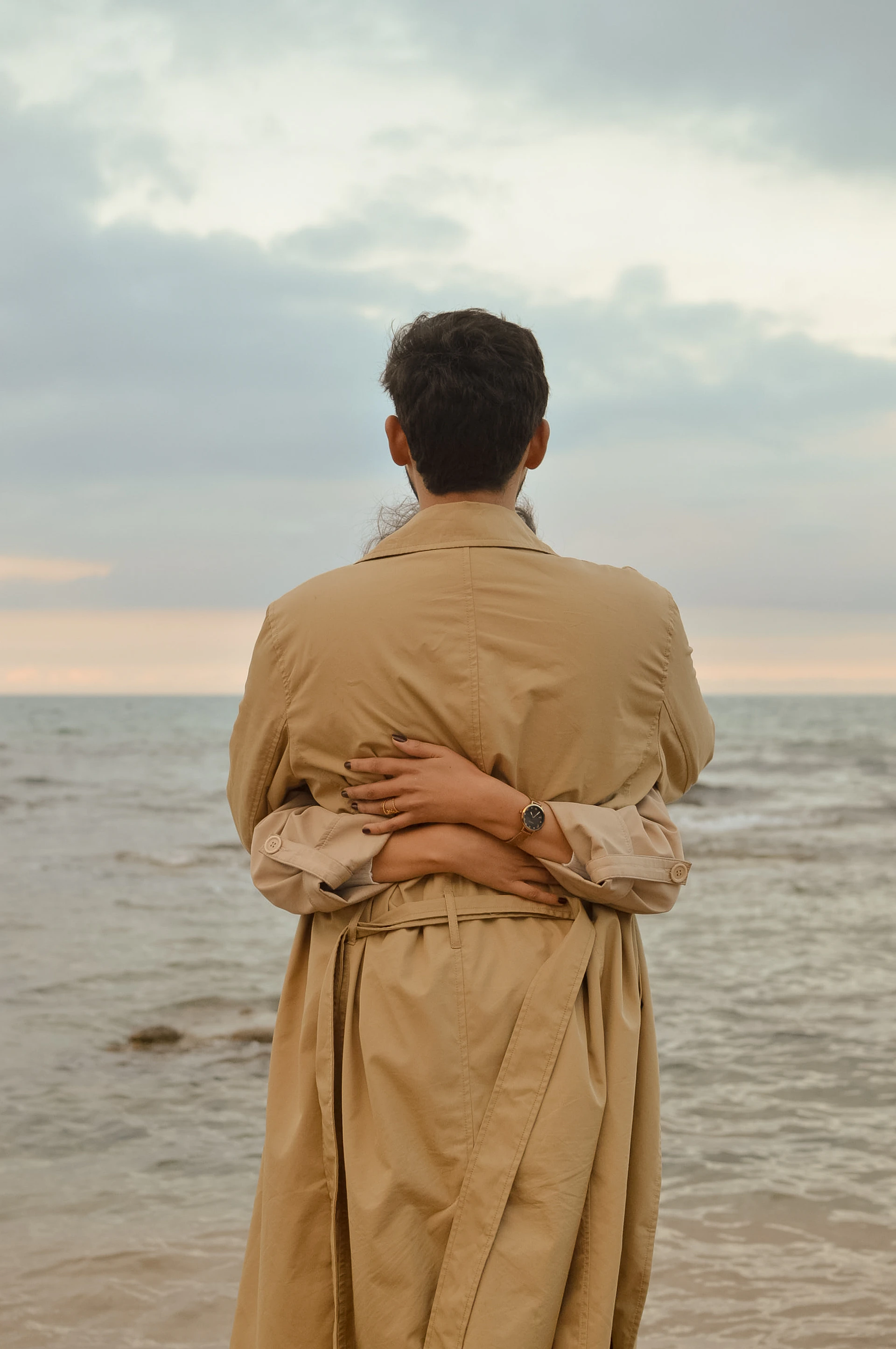 Person in trench coat embraced by another by the ocean