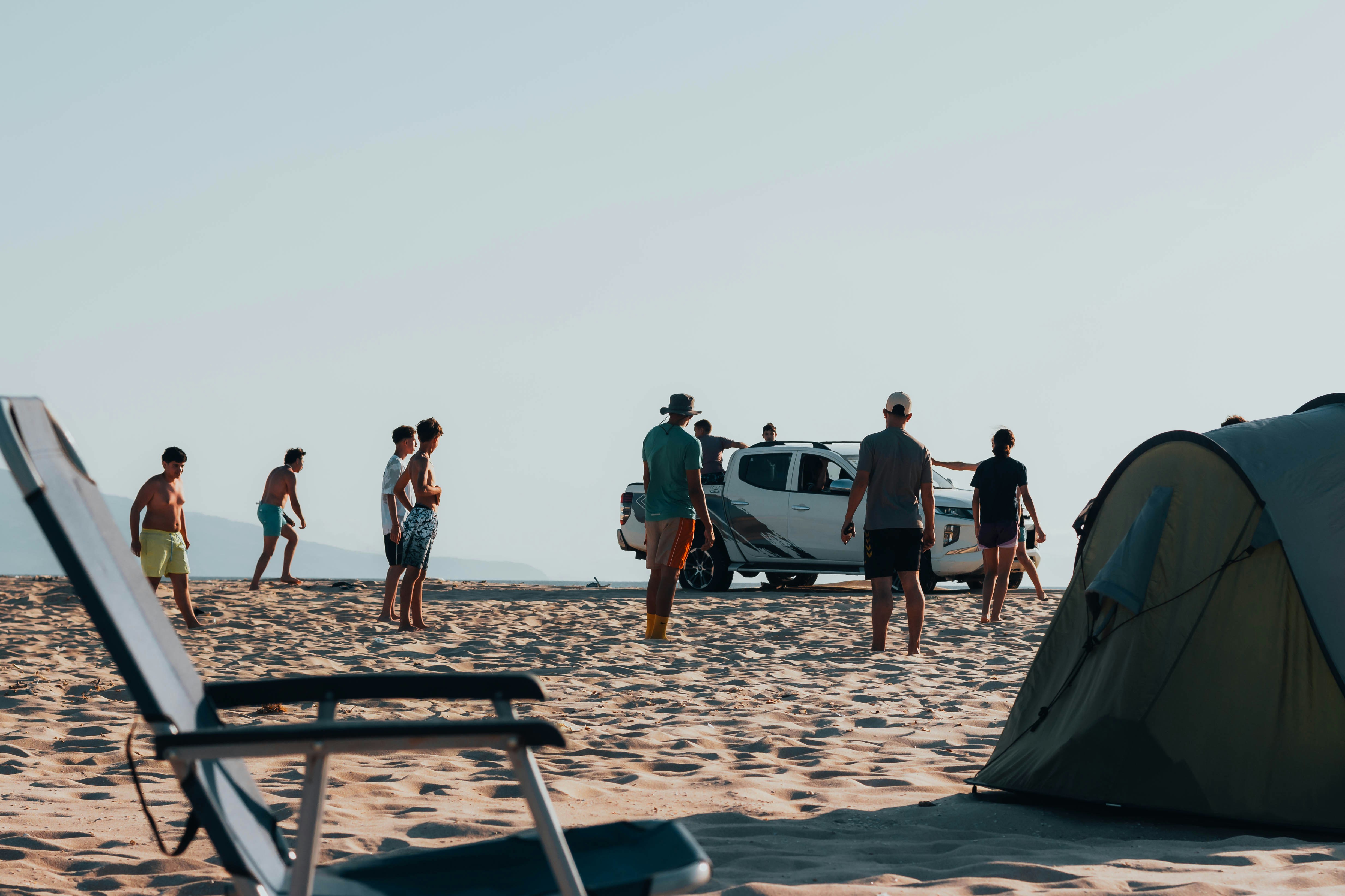 Escena de playa con personas, tienda de campaña, silla y vehículo.