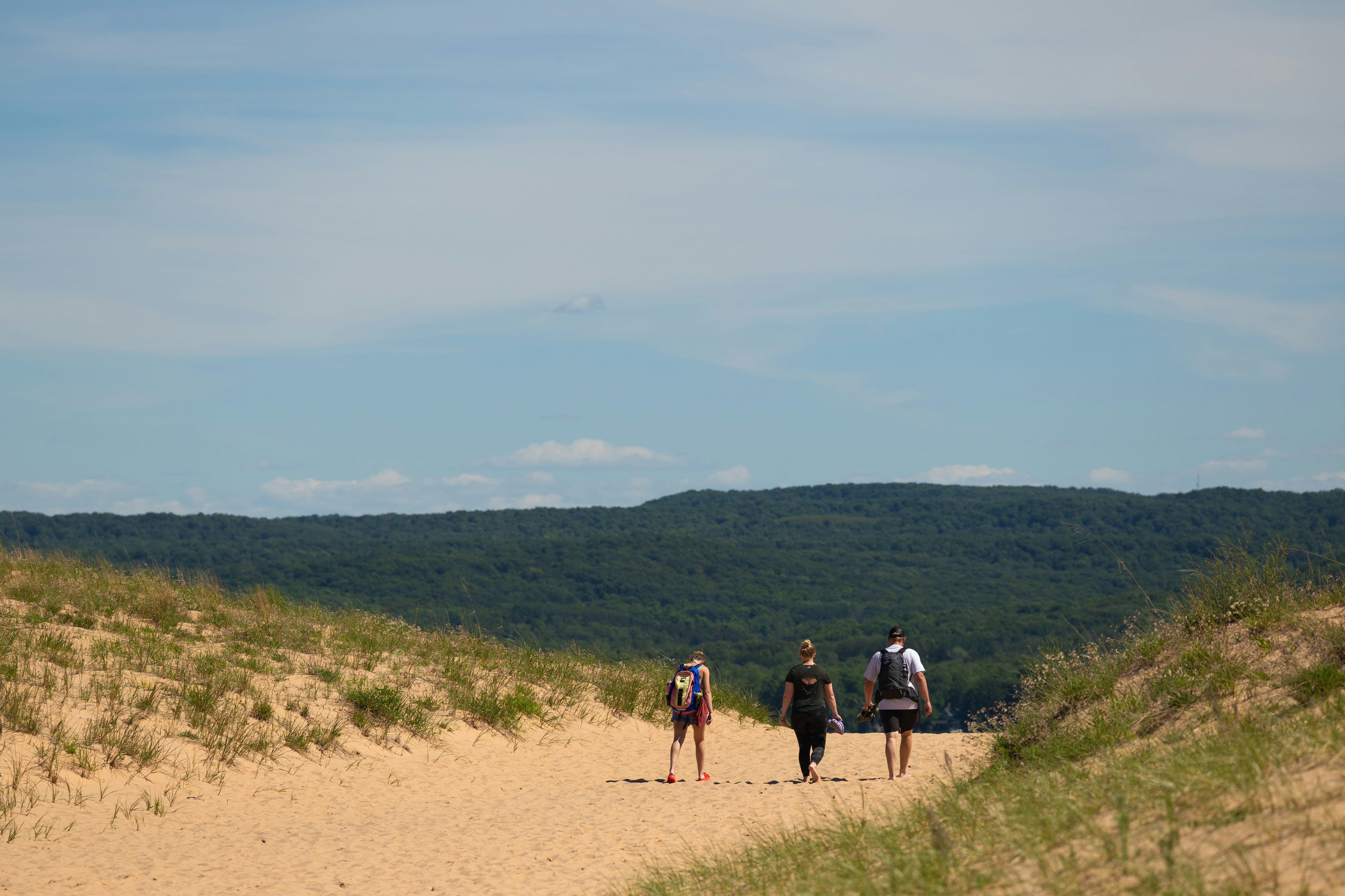 Tre persone camminano su un sentiero sabbioso tra dune erbose.