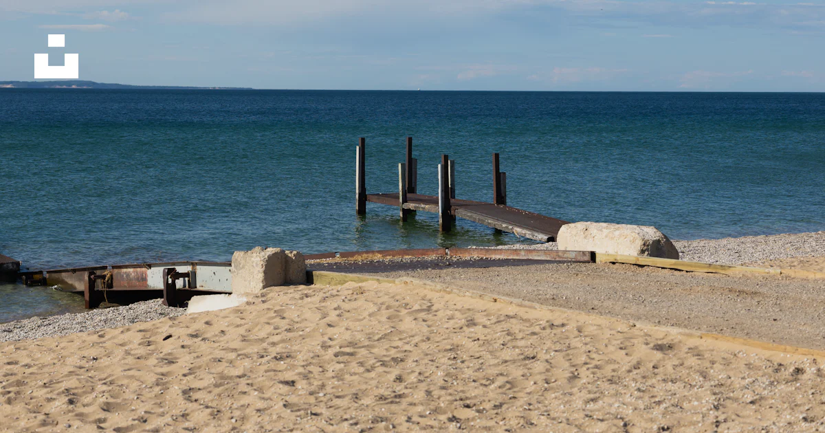 Stack Lakeshore Playa de arena con un muelle de madera que se adentra en aguas azules ...