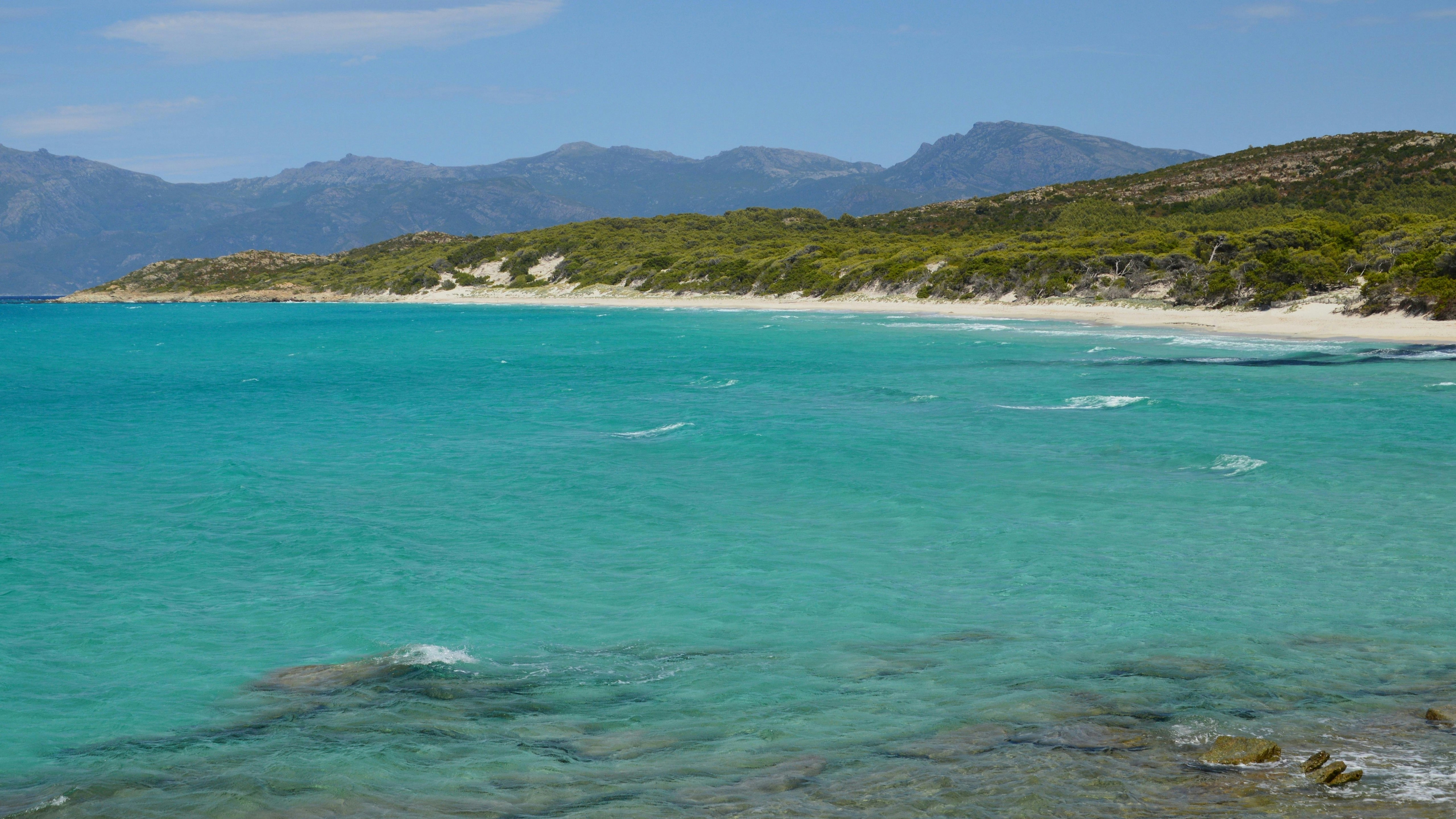 Océan turquoise clair, plage blanche, collines verdoyantes et montagnes lointaines.