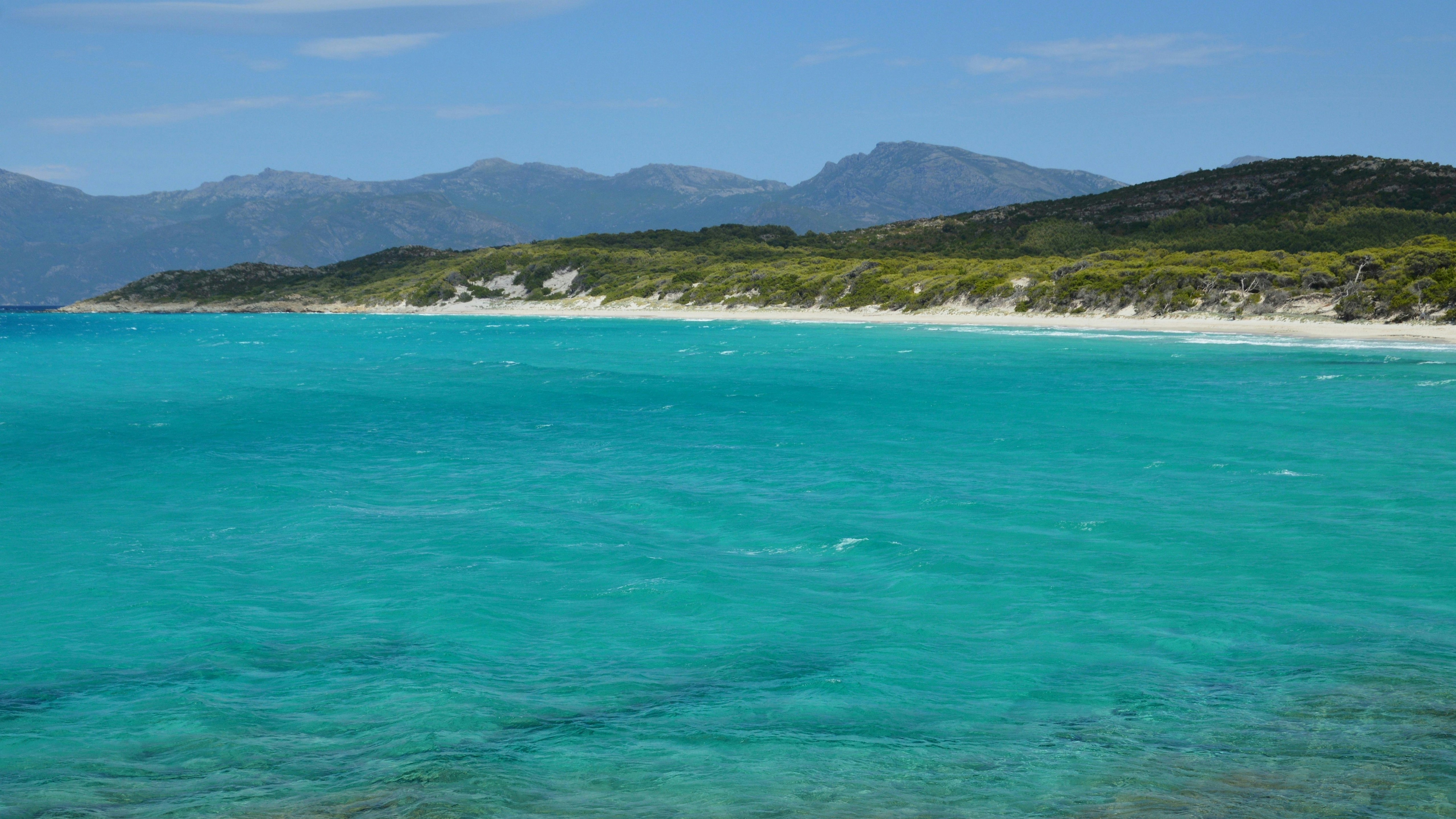 Eau turquoise claire, plage de sable blanc et montagnes lointaines.