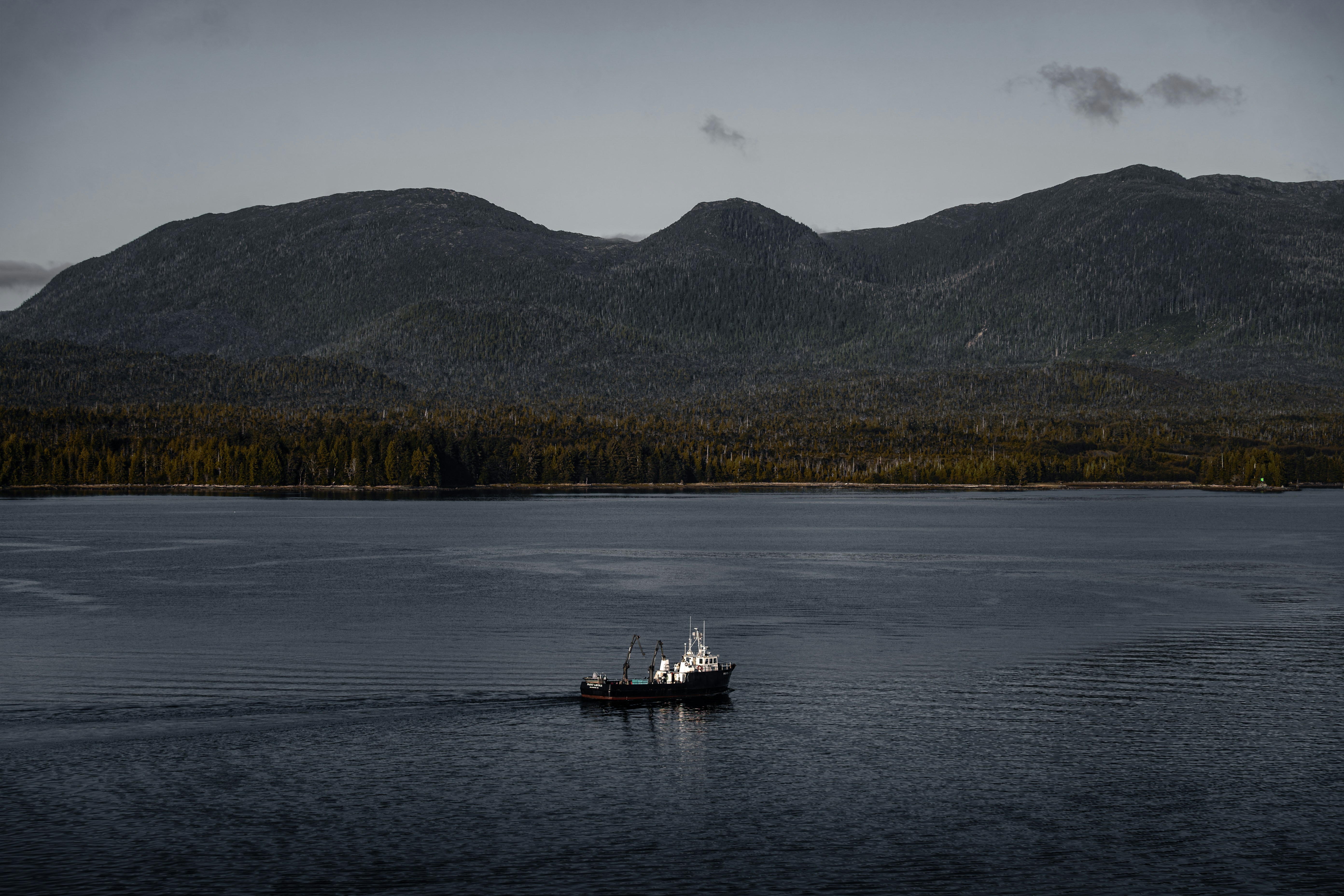 Un bateau navigue dans des eaux calmes avec des montagnes sombres et boisées derrière.