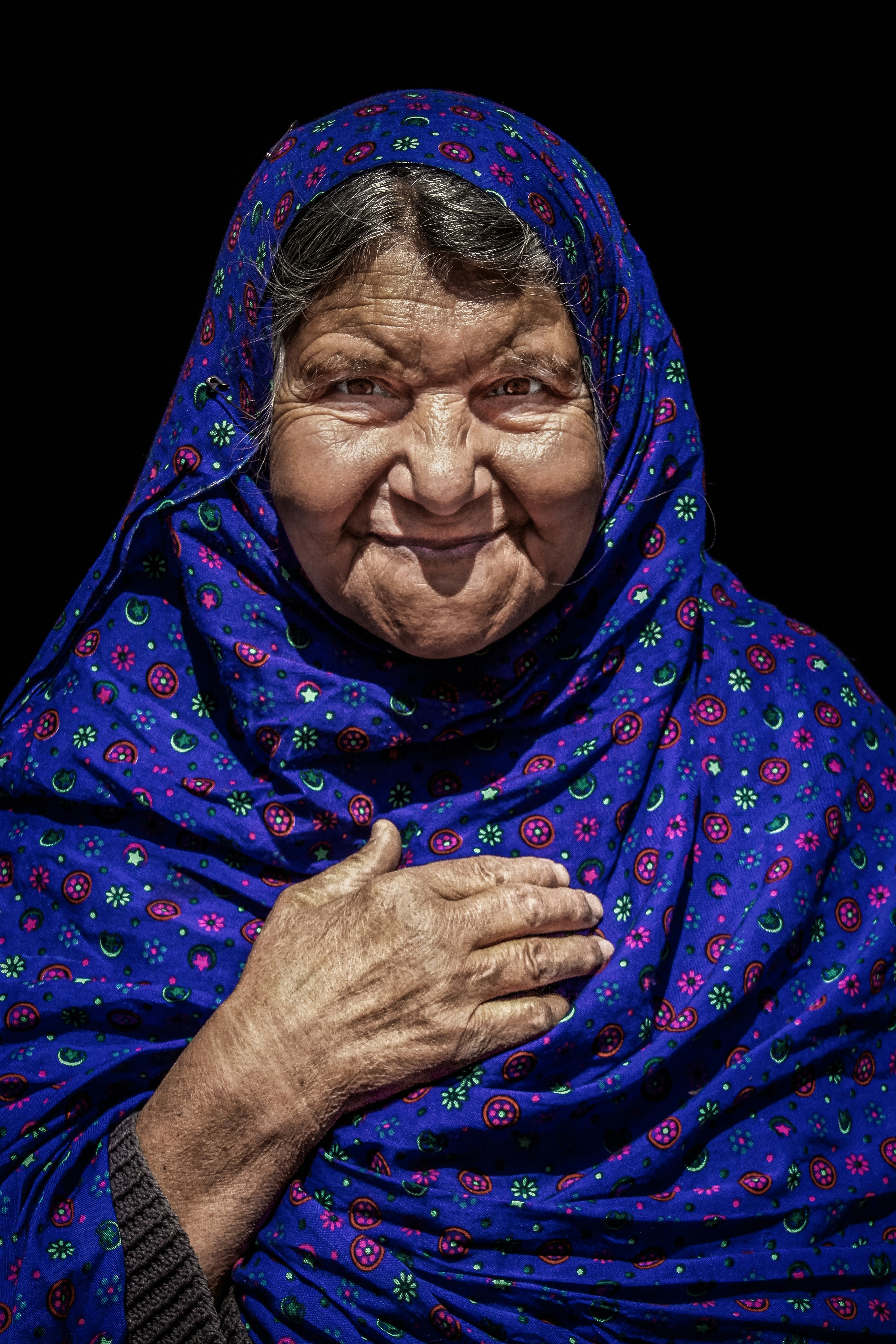 Elderly woman in blue headscarf smiling with hand on chest.