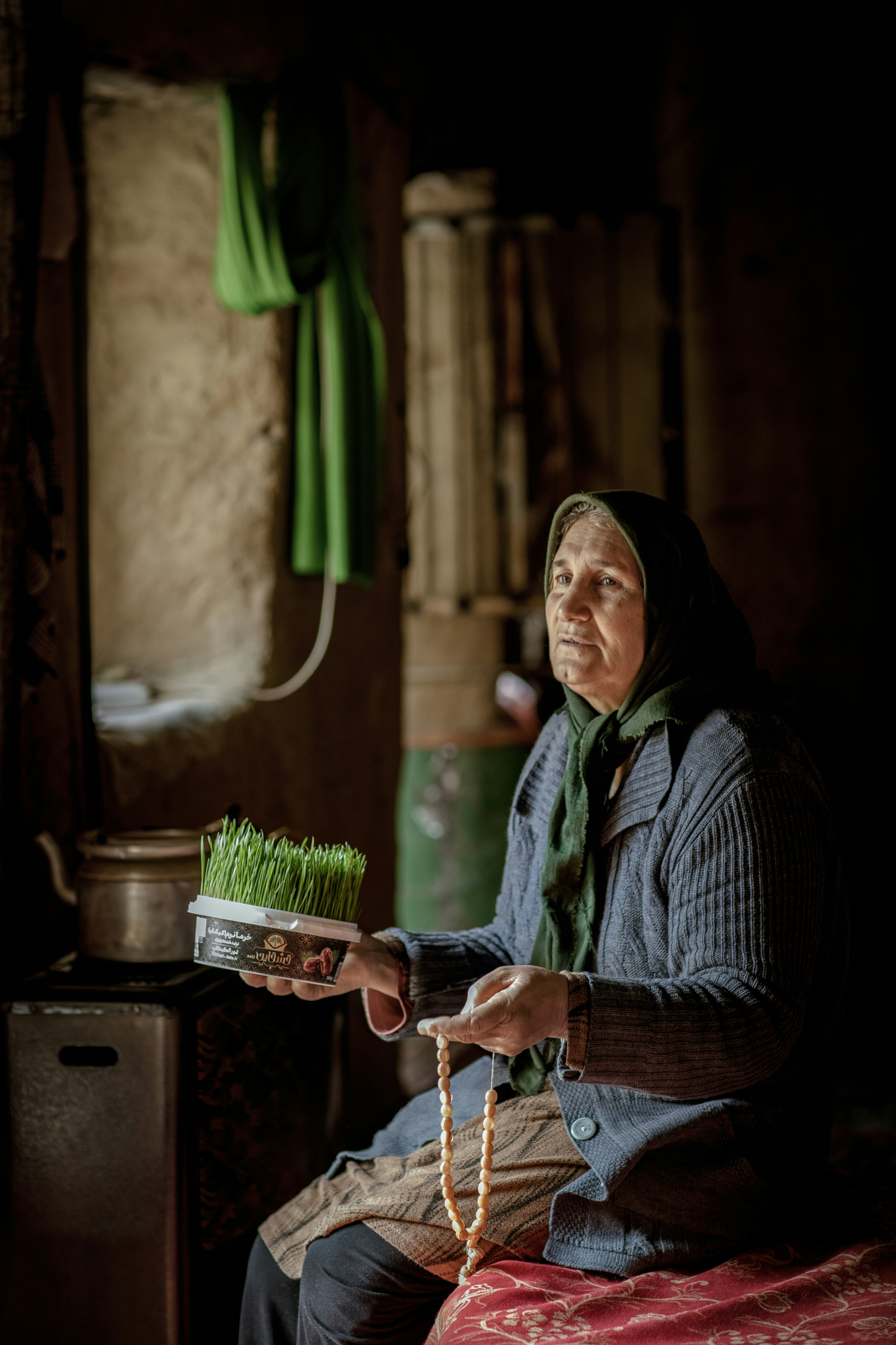 An elderly woman holds green sprouts and prayer beads.