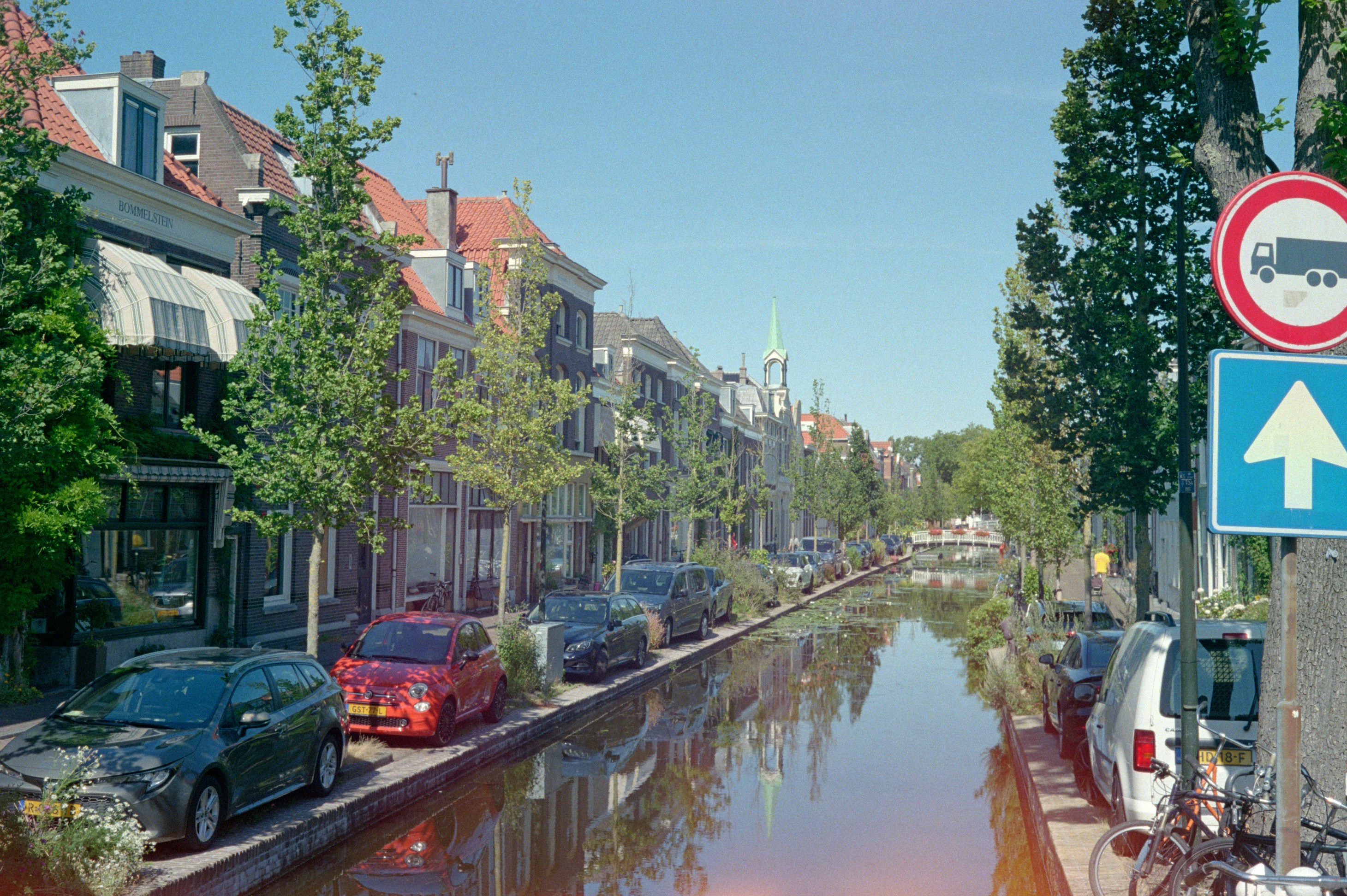 A sunny canal scene with buildings, parked cars, and trees.
