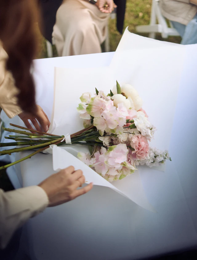 Mani che avvolgono nella carta bianca un mazzo di ortensie e garofani rosa