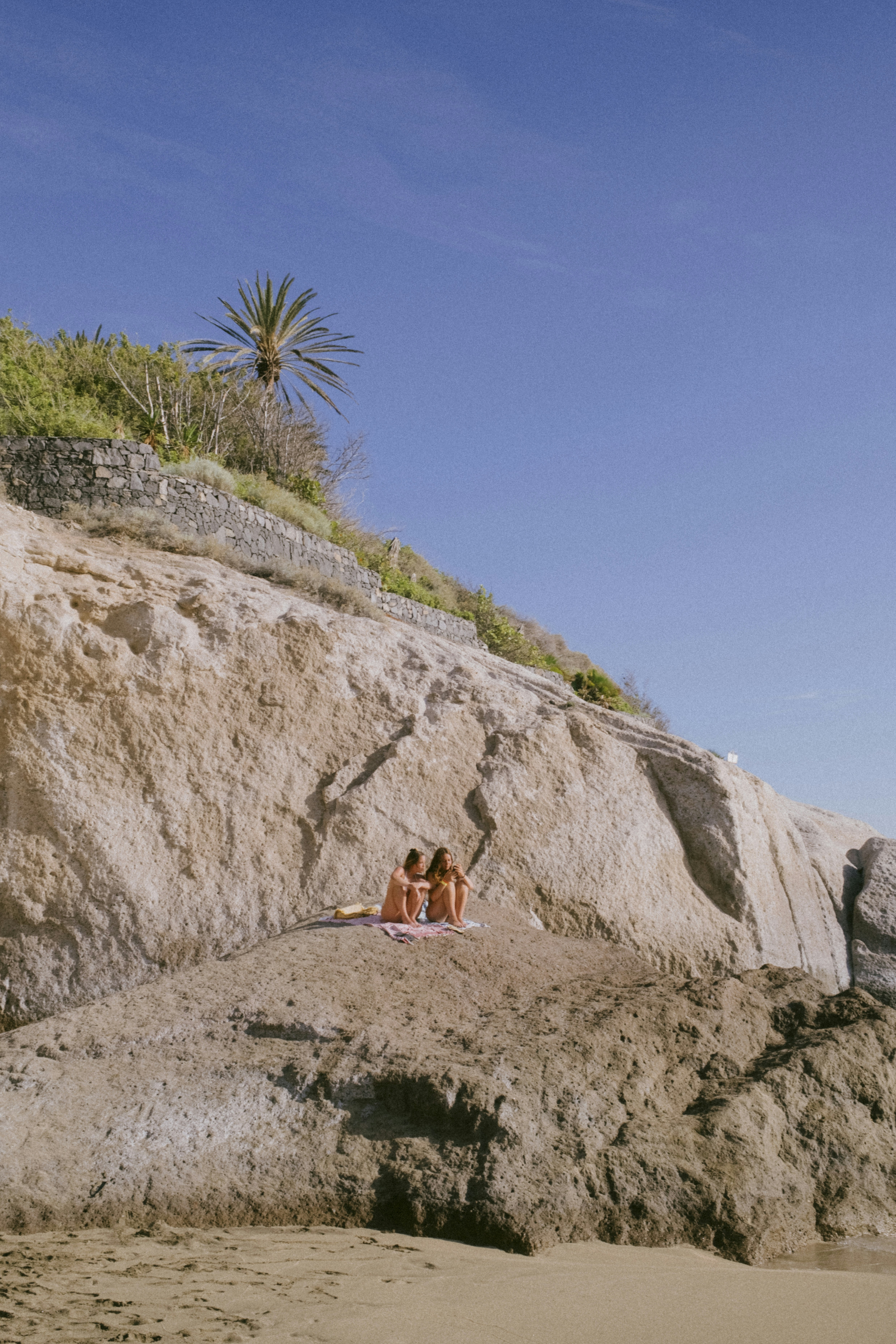 Two people relaxing on a rocky beach below a cliff.