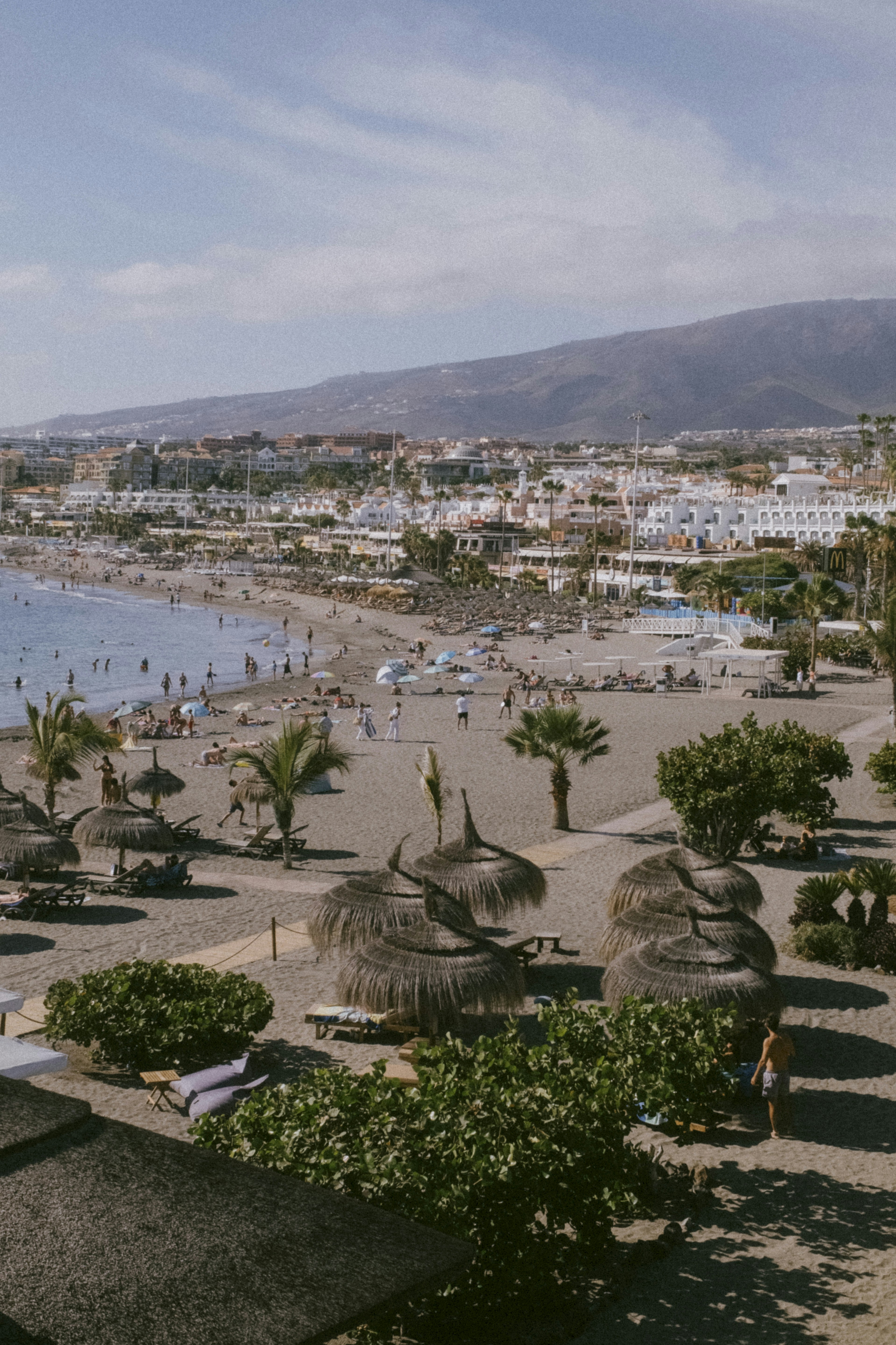 Sunny beach with people, straw umbrellas, and a coastal town.