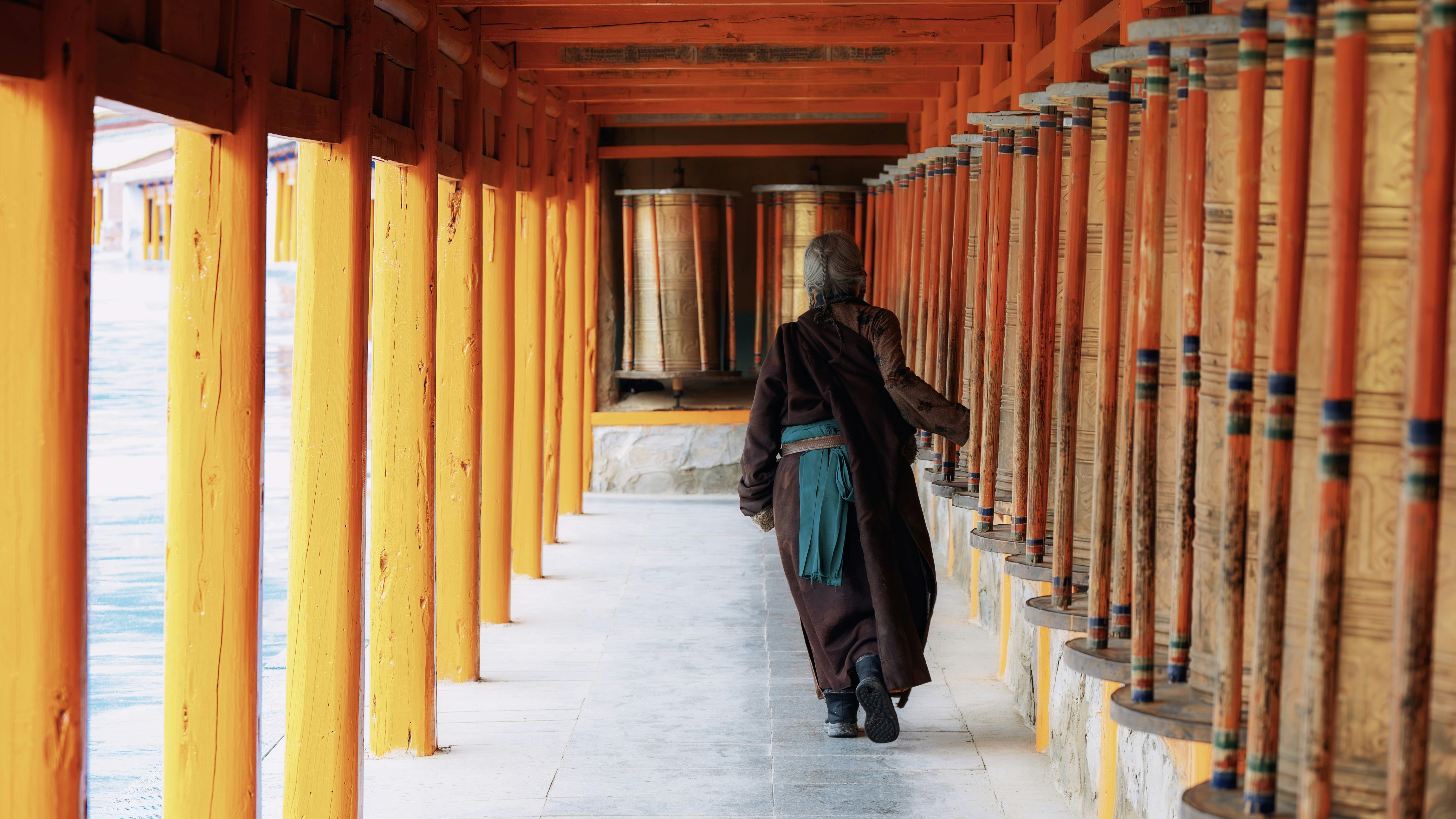 Monk walking past prayer wheels in a temple corridor.