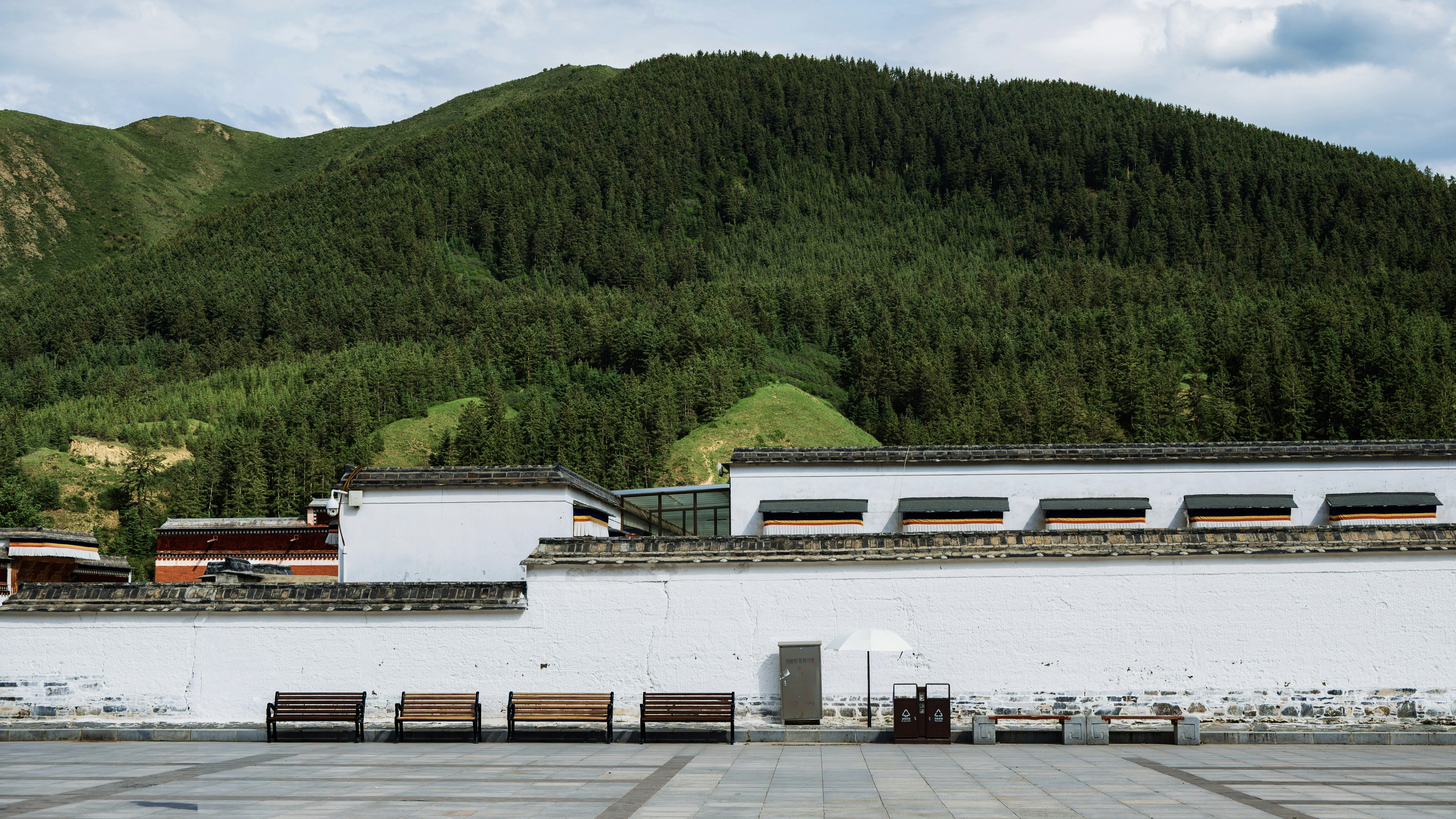 White wall with benches in front of a forested mountain.