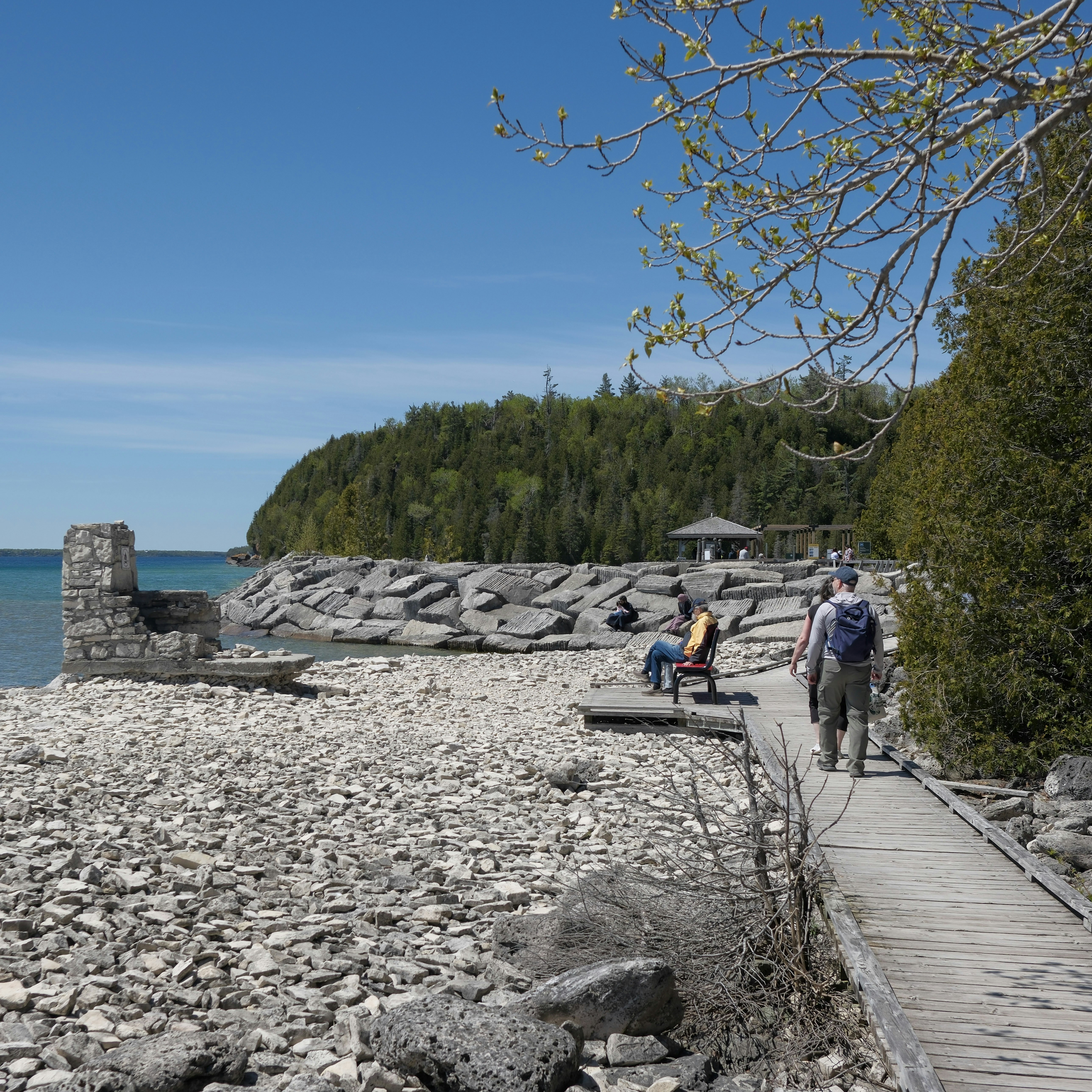 People walk on boardwalk by rocky beach and blue water.