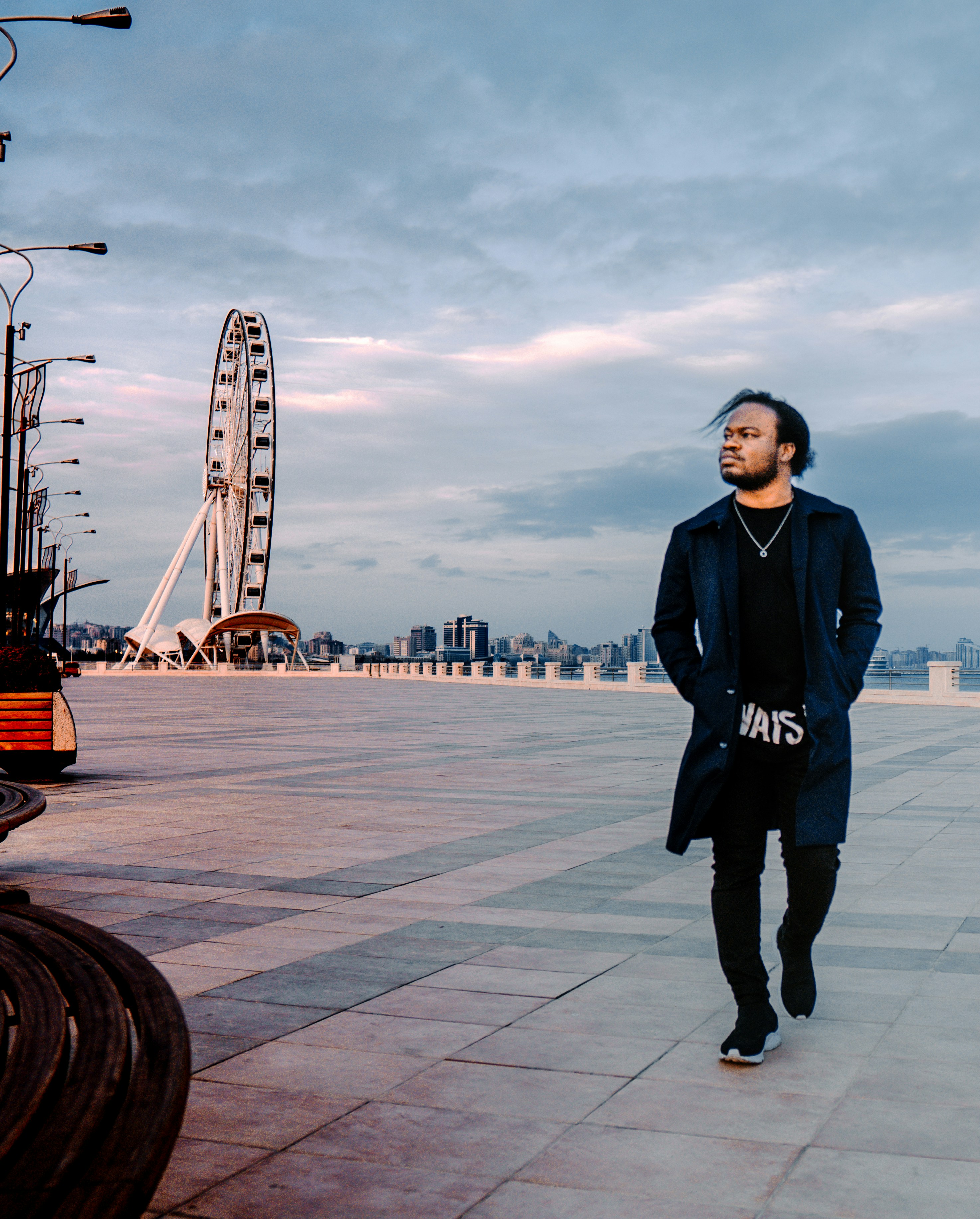Man walks on promenade with ferris wheel under cloudy sky.