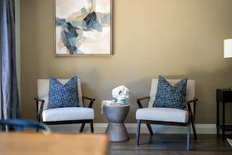 Modern armchairs with pillows, side table, and abstract art.